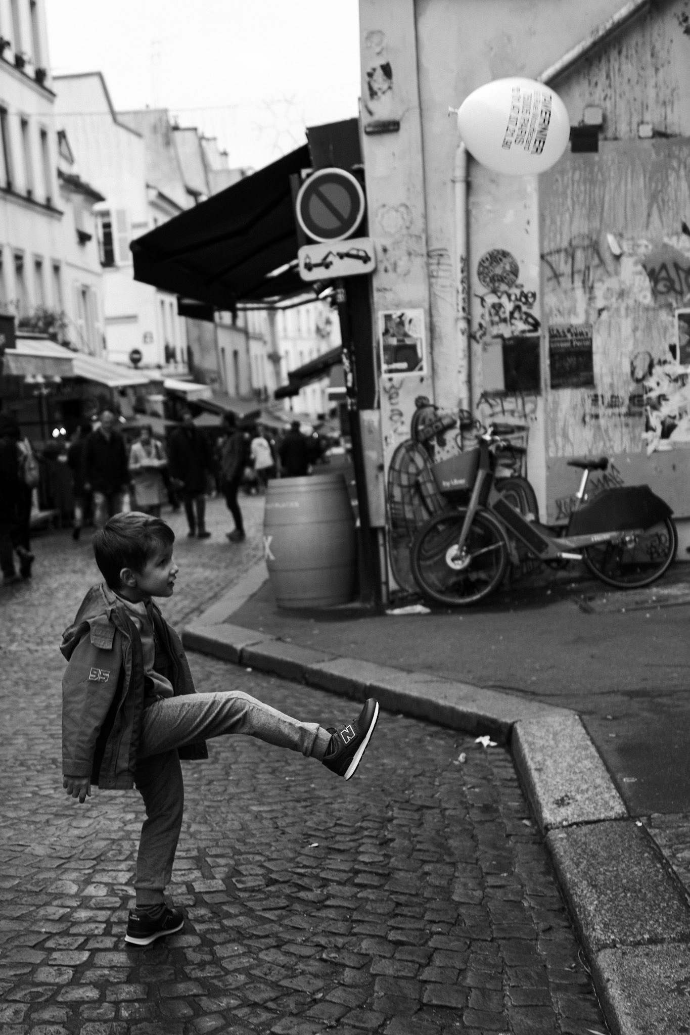 Un jeune garçon lève la jambe comme pour shooter dans un ballon invisible, dans une rue pavée animée de Montmartre ; un ballon blanc marqué « Happy Birthday » flotte à proximité, suspendu dans l’air urbain.
