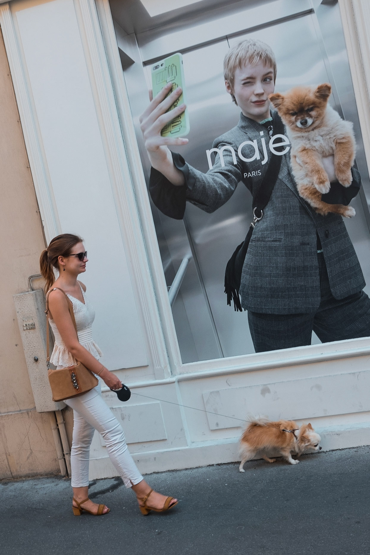 Une femme promenant un petit chien passe devant une grande affiche de mode représentant une autre femme tenant un chien similaire, créant une mise en abîme visuelle.