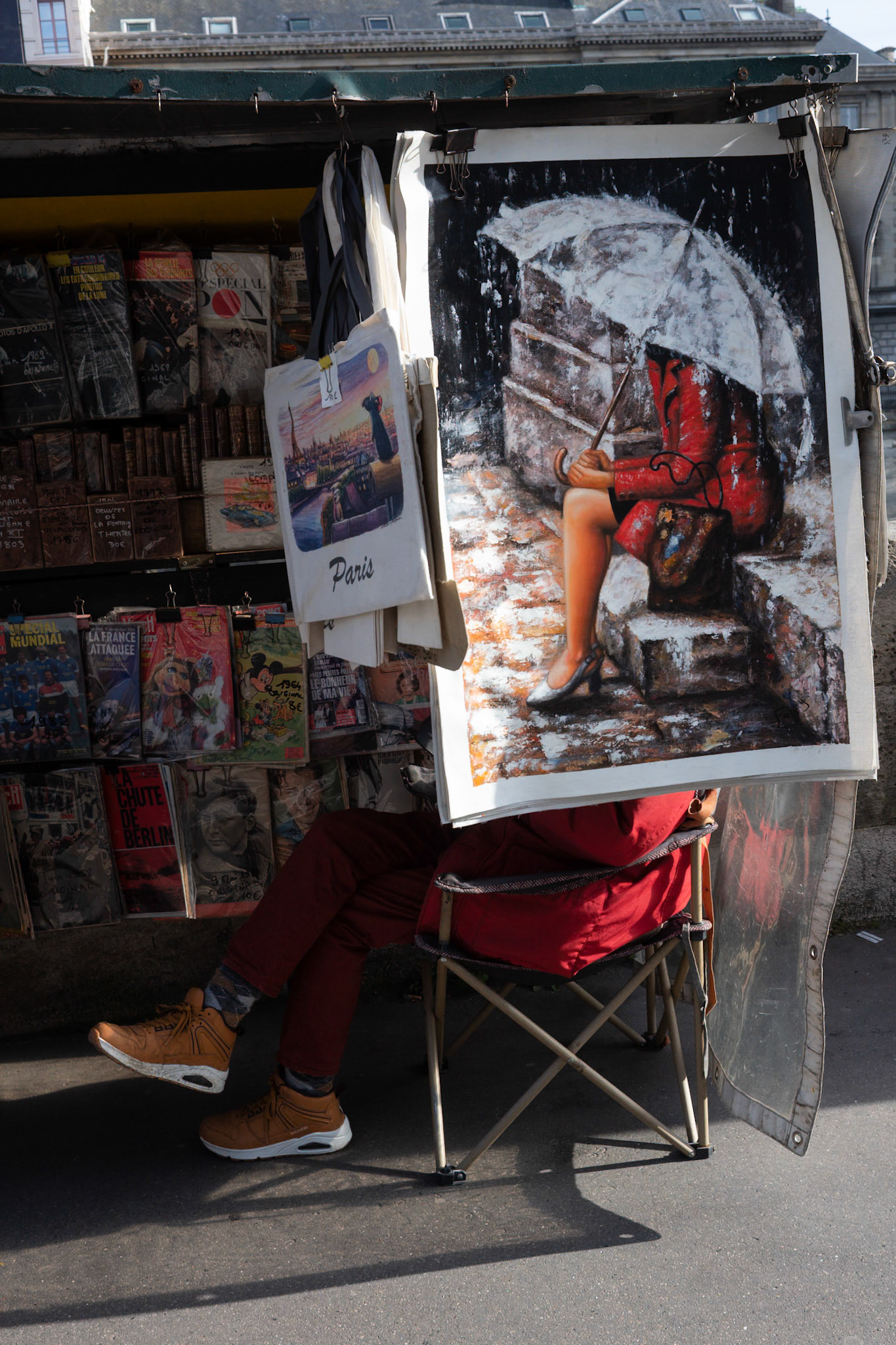 Un bouquiniste parisien assis derrière une grande peinture représentant une femme en rouge sous un parapluie, son corps se confondant avec l’illustration.