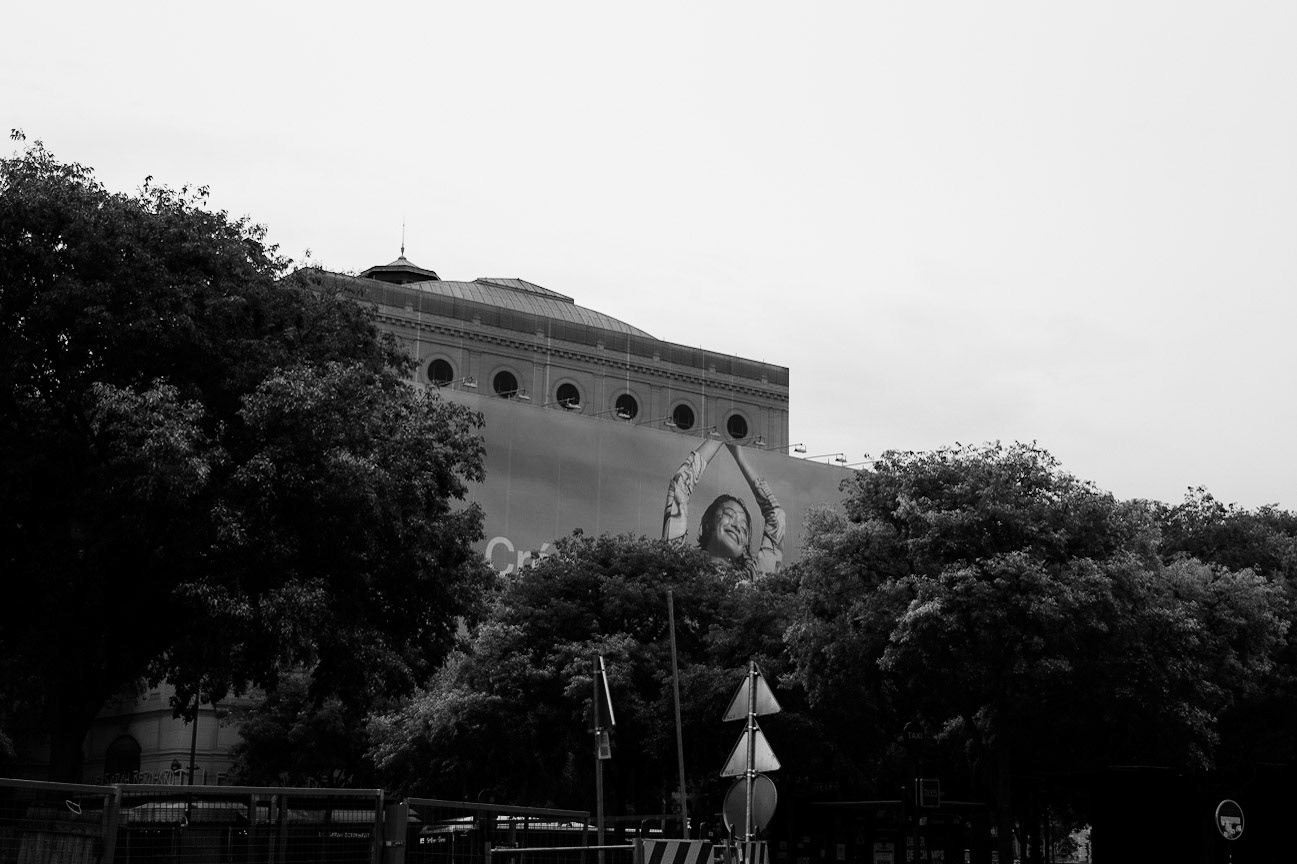 Une immense affiche publicitaire représentant une femme souriante avec les bras levés dépasse des arbres devant une façade monumentale, dans une composition mêlant nature urbaine et image surdimensionnée.