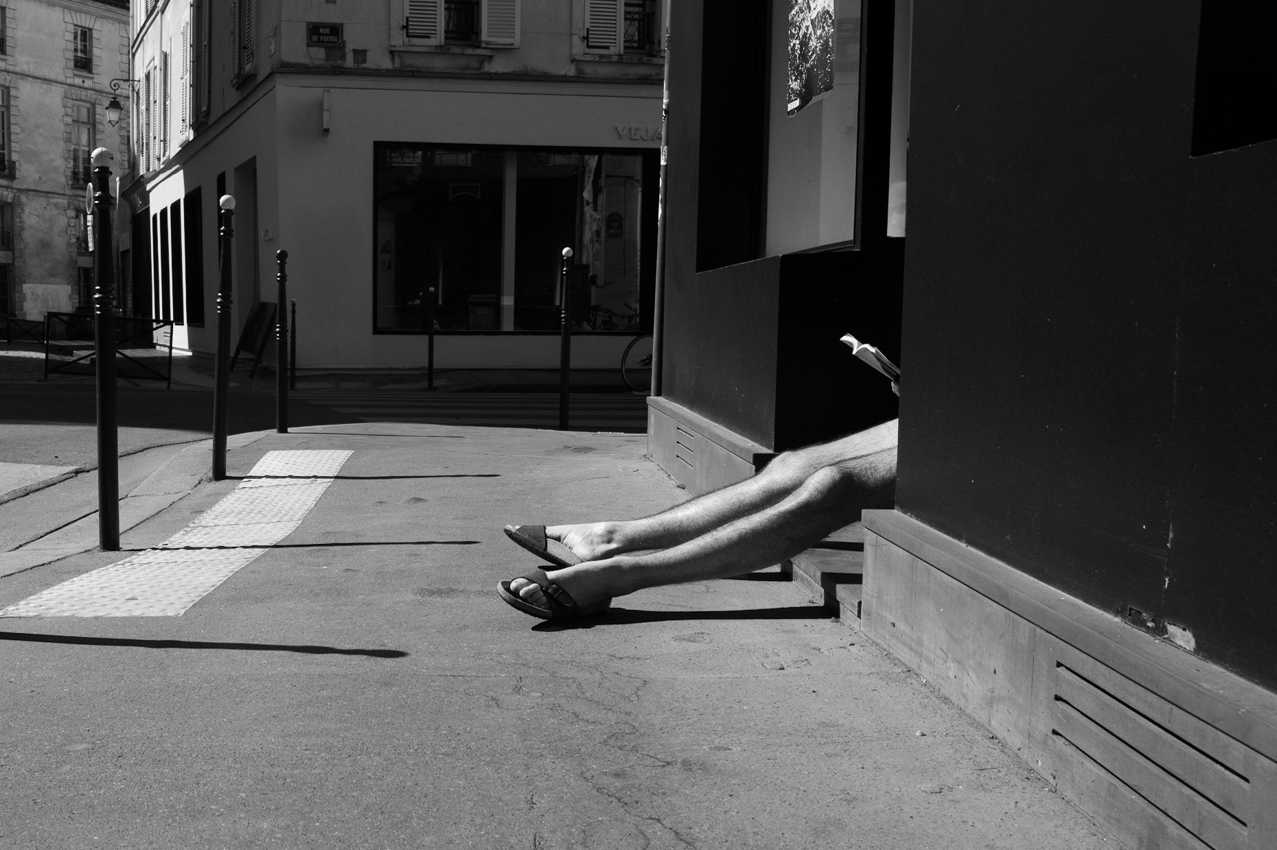Un homme assis à l’ombre d’un mur noir, jambes allongées au soleil, lisant un livre, le reste de son corps caché par l’angle du bâtiment.