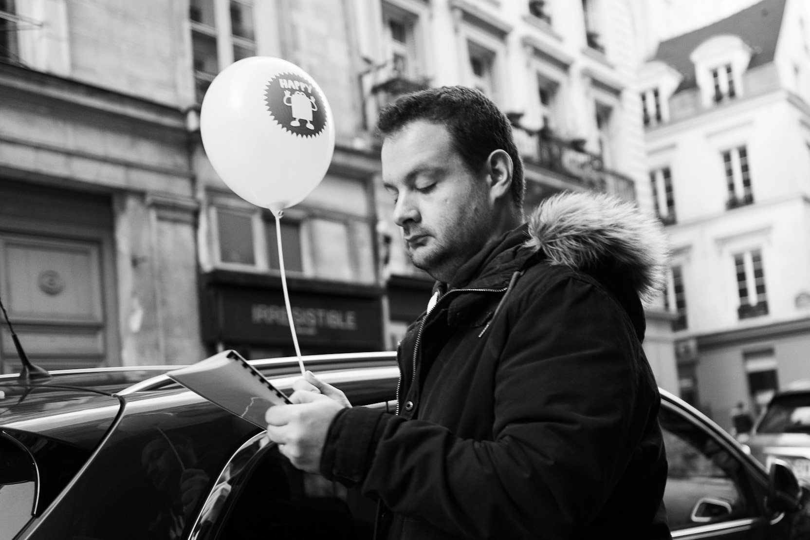 Un homme concentré lit un carnet, debout près d’une voiture dans une rue parisienne. Un ballon « Happy » flotte au-dessus de lui, comme en décalage avec son expression sérieuse.