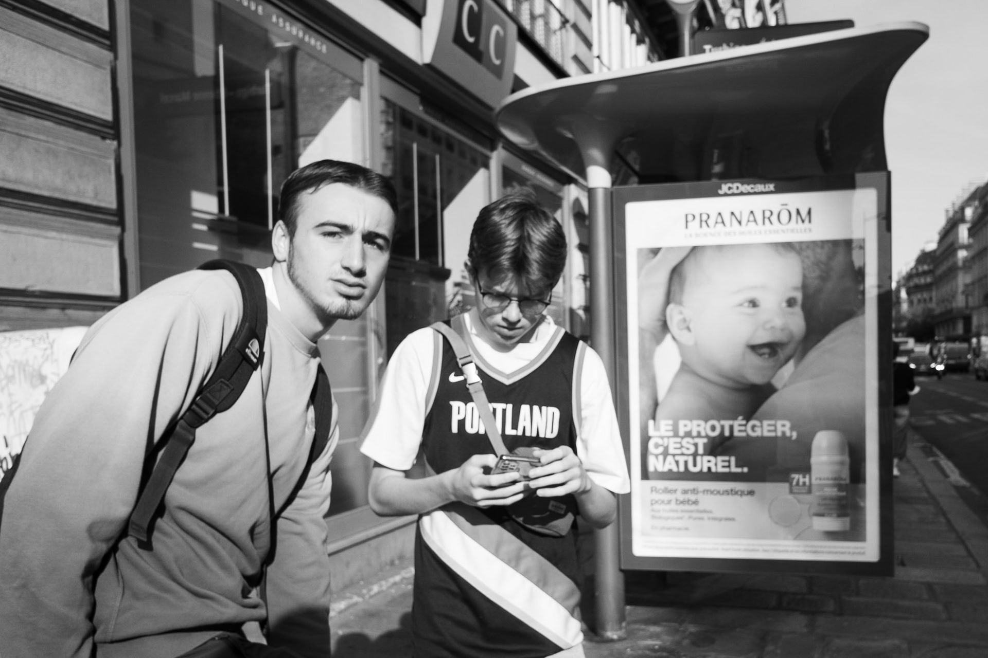 Deux adolescents sur un trottoir parisien, l’un regardant l’objectif, l’autre concentré sur son téléphone. À l’arrière-plan, une publicité avec un bébé souriant.