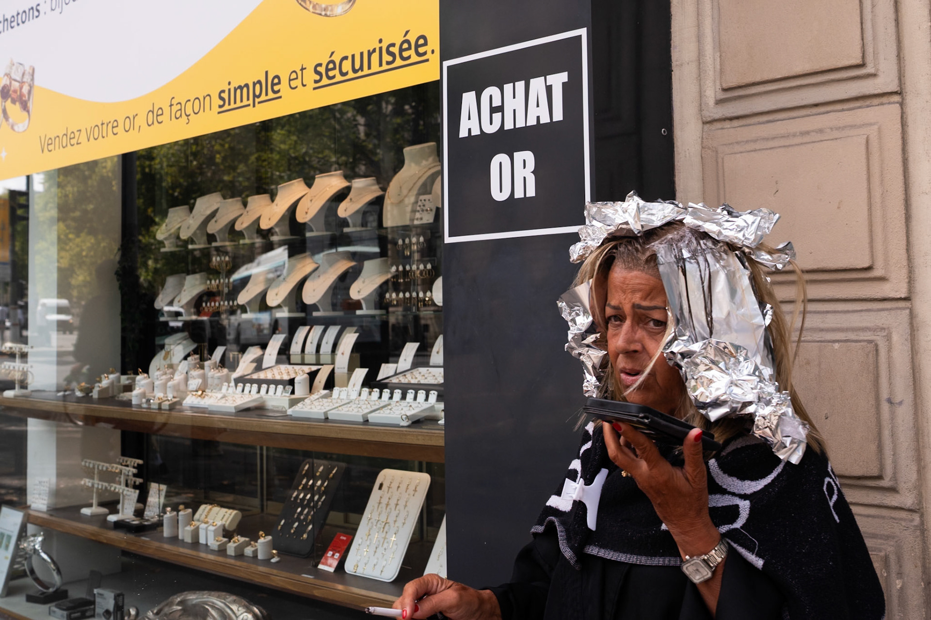 Une femme à la mise en plis recouverte de papiers d’aluminium fume une cigarette devant une vitrine de bijouterie, sur fond d’affiche "Achat Or", dans un contraste ironique entre glamour brut et luxe exposé.