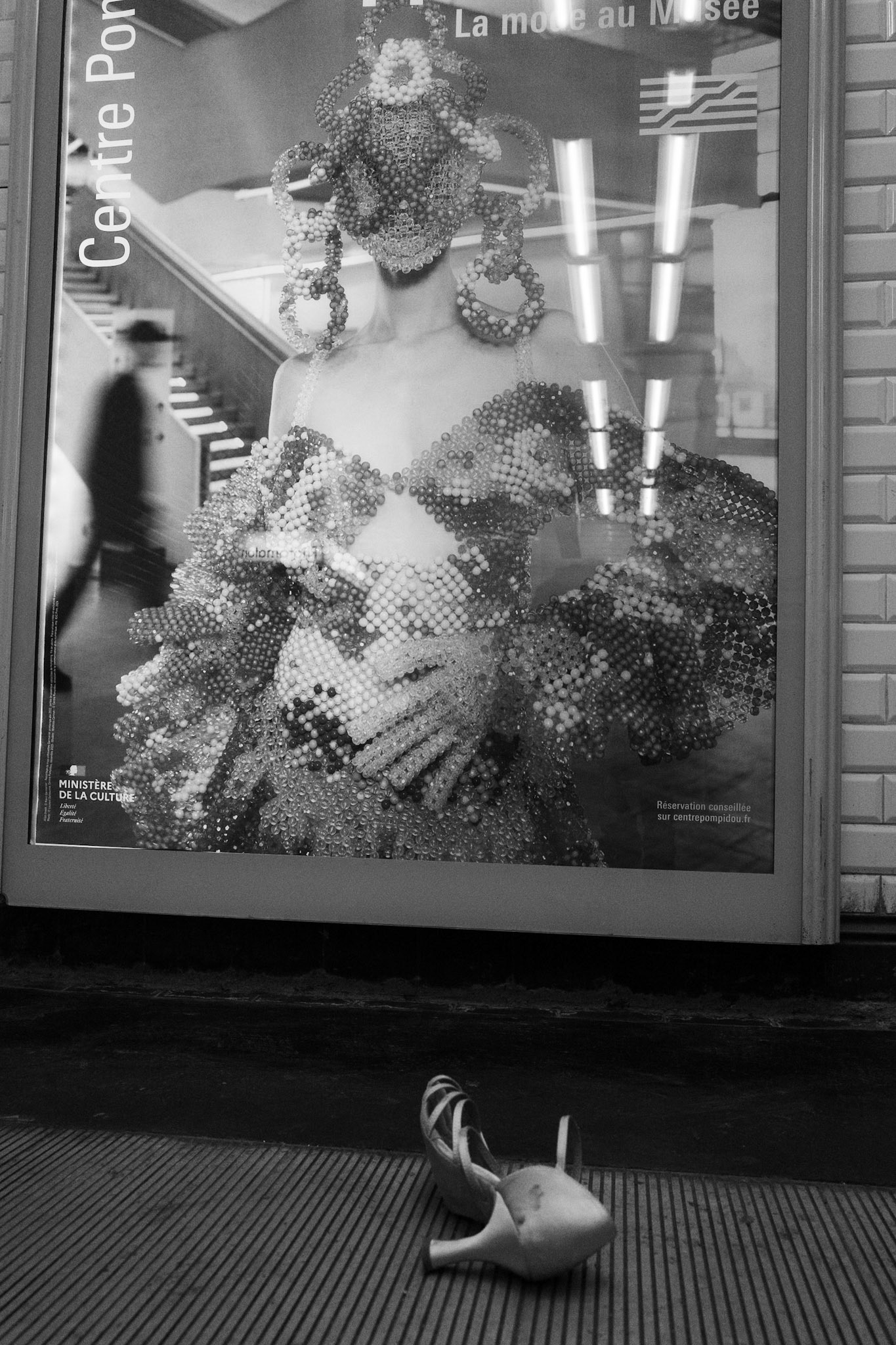 Une chaussure à talon solitaire abandonnée au sol dans une station de métro, devant une affiche de mode aux motifs baroques et perles étincelantes.