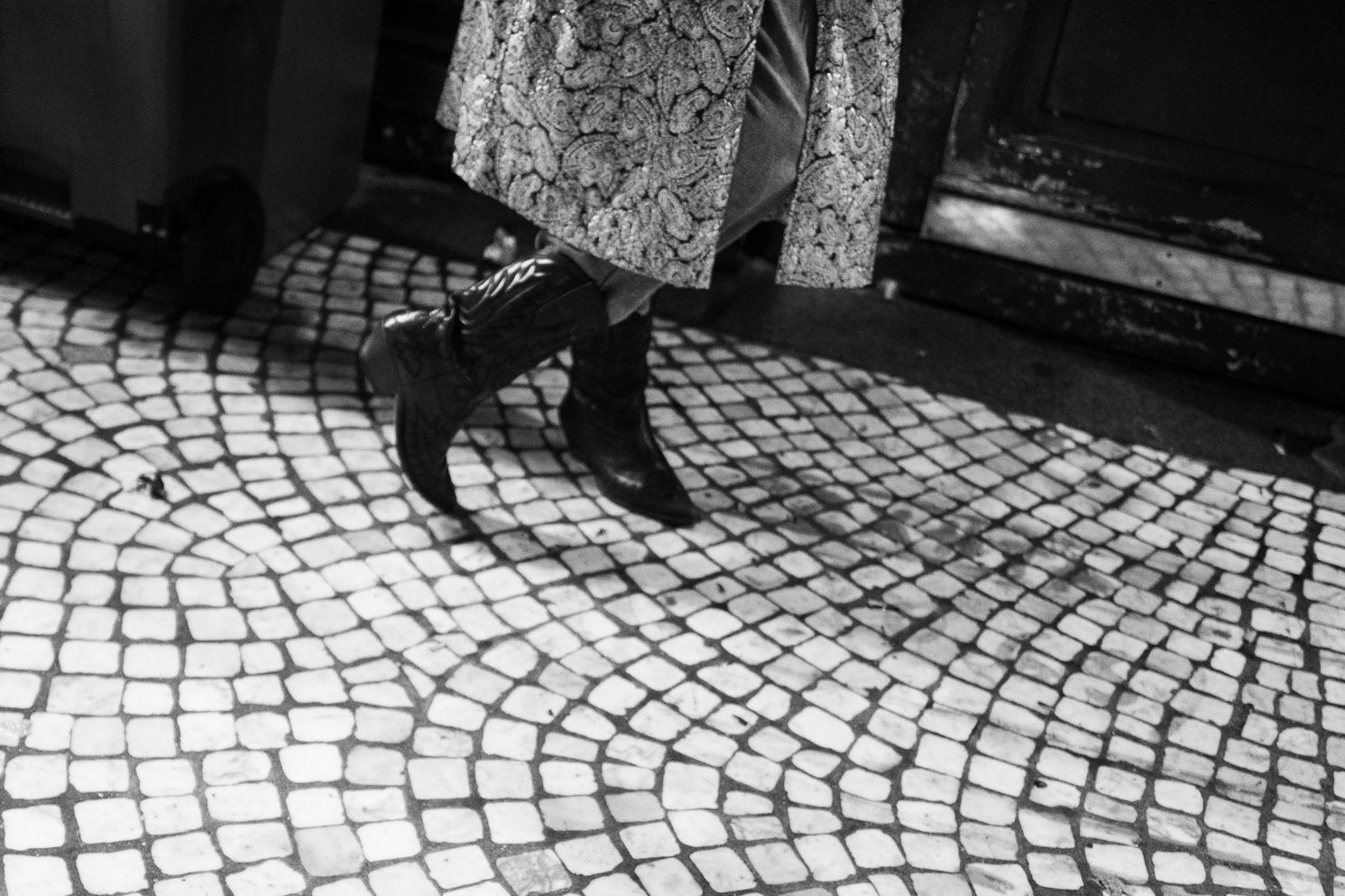 Gros plan sur les jambes croisées d’une femme en bottes et manteau brodé, marchant sur un sol en pavés arrondis — une scène anonyme et graphique au ras du trottoir.