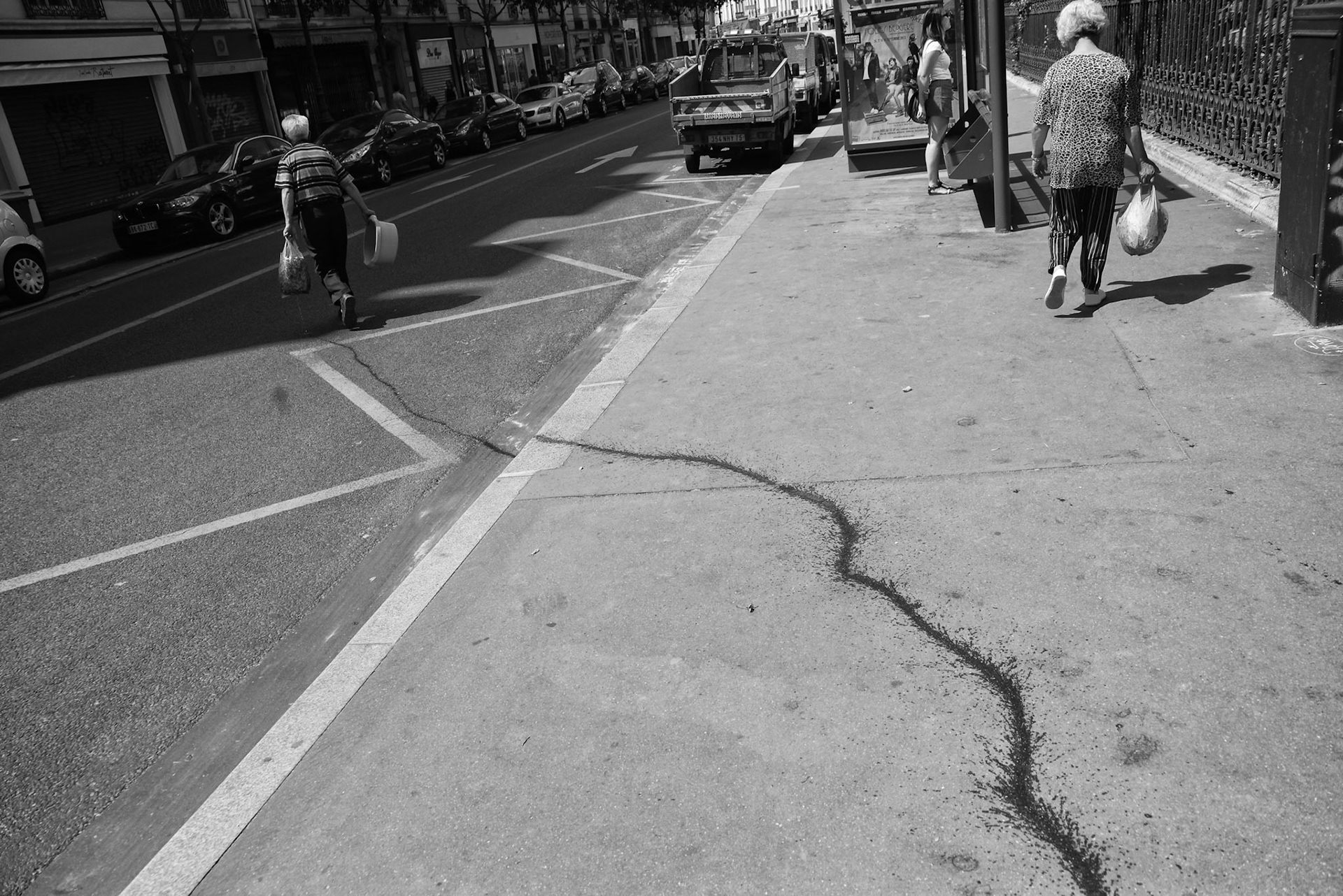 Deux personnes âgées marchent en sens opposé sur un trottoir marqué d’une longue traînée noire en zigzag, accentuant la géométrie urbaine.