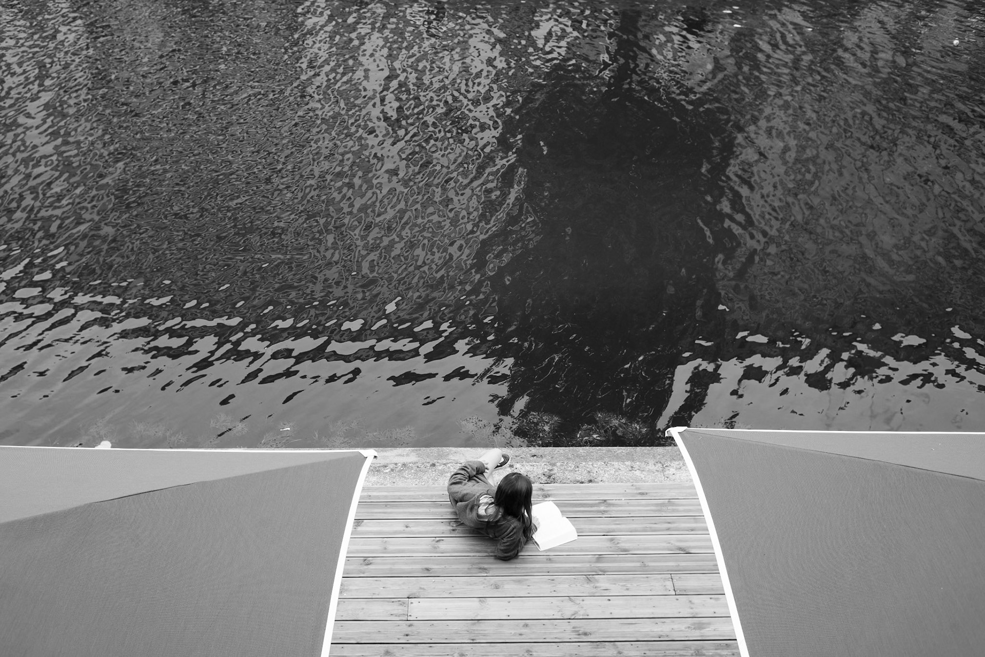 Une jeune femme assise seule sur une terrasse en bois au bord de l'eau, lisant un livre, vue du dessus. La surface de l'eau reflète des formes abstraites et ondulées.