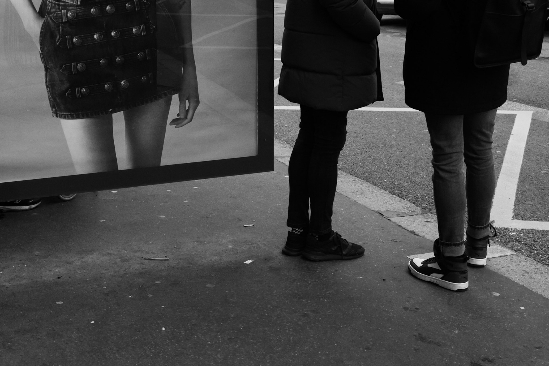 Scène de rue capturant les jambes de deux passants en face d'une publicité affichant les jambes d'une femme, créant un jeu visuel entre réel et image.
