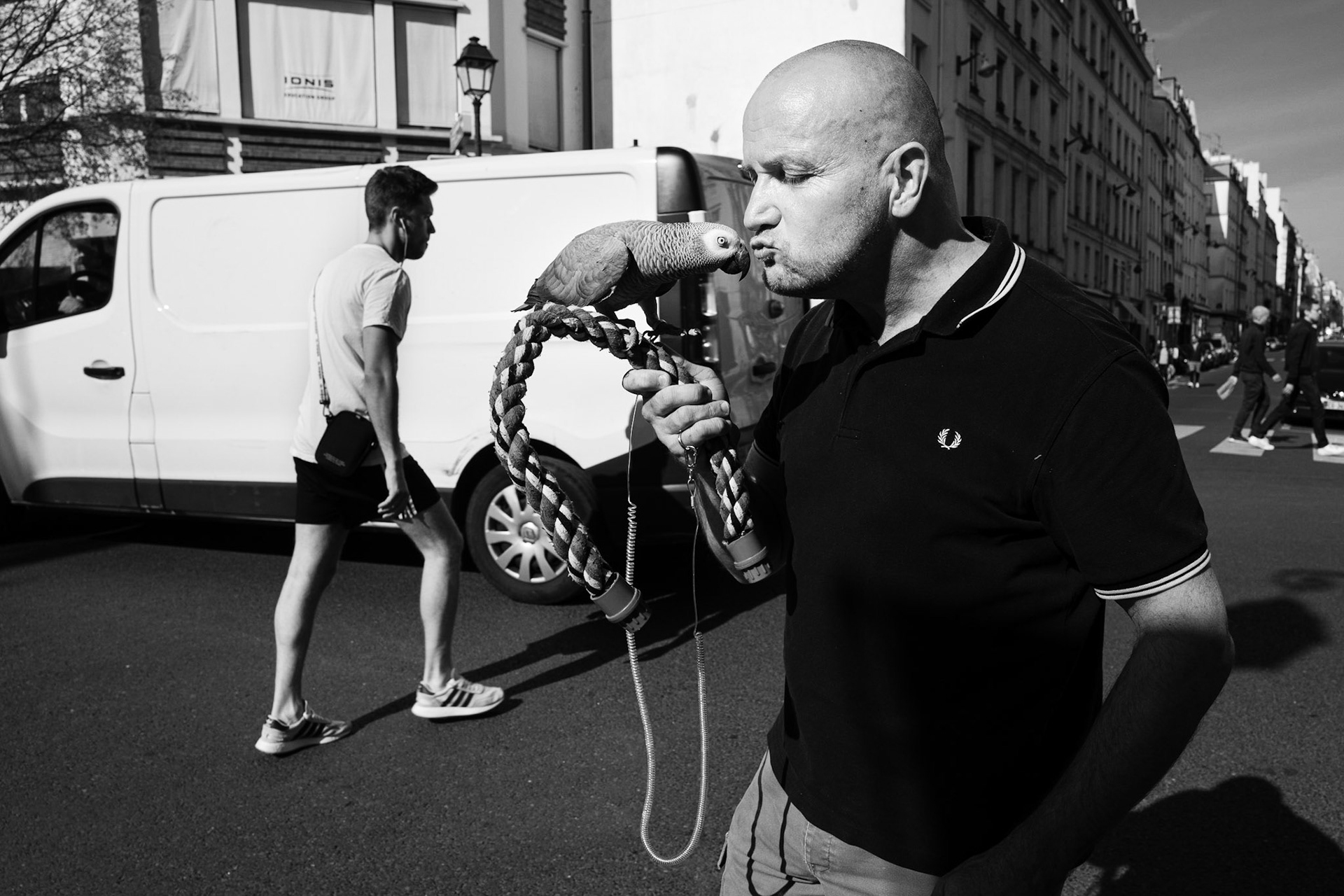 Un homme embrasse affectueusement un perroquet perché sur une corde dans une rue parisienne, avec la vie urbaine en arrière-plan.