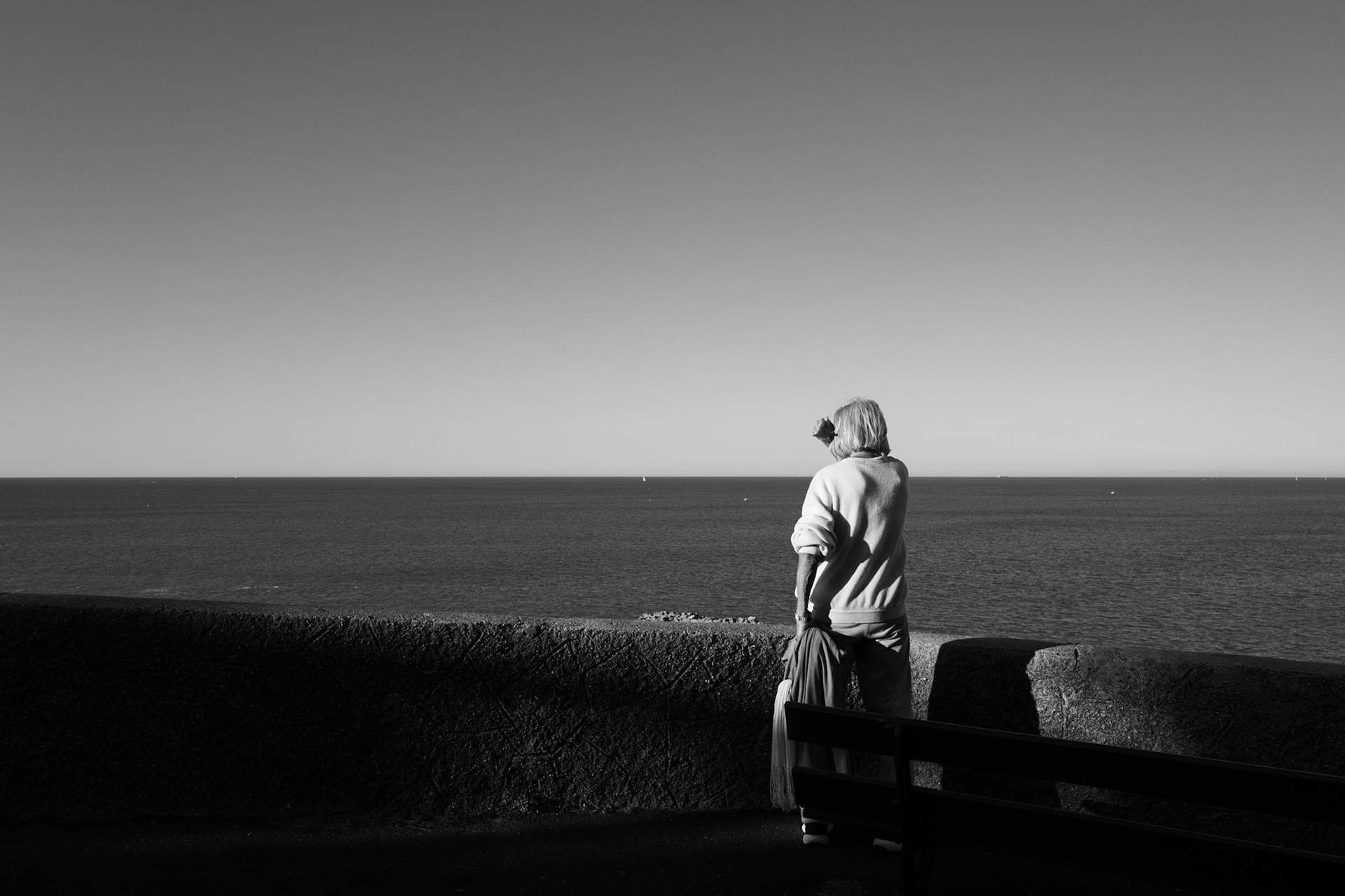 Une femme âgée, vue de dos, regarde l’horizon sur une promenade en bord de mer, seule face à l’immensité de l’eau et du ciel.