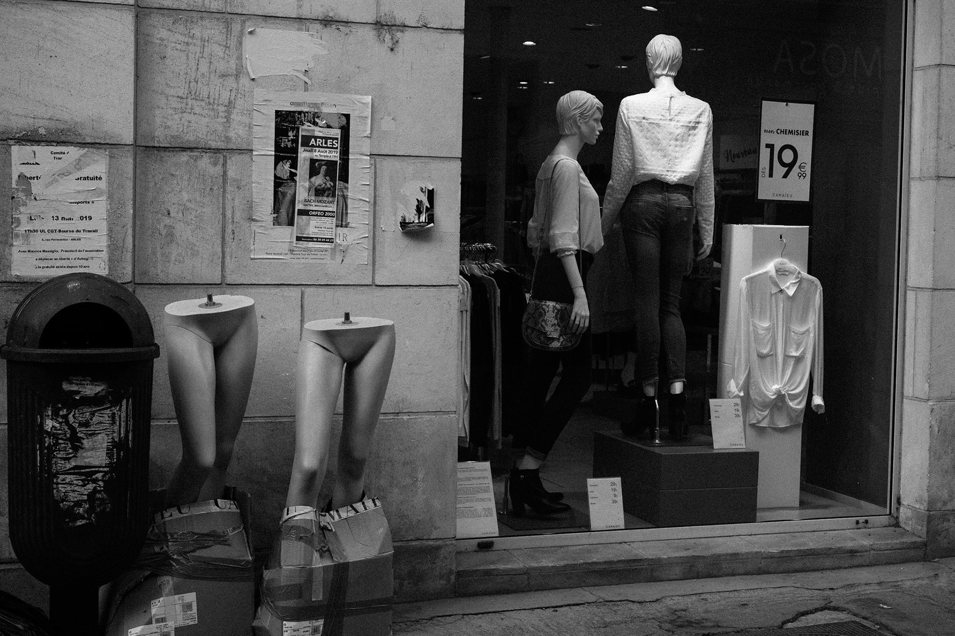 Devanture de boutique avec des mannequins en vitrine et deux jambes de mannequin sans buste posées au sol, attachées avec du ruban adhésif, à côté d'une poubelle.