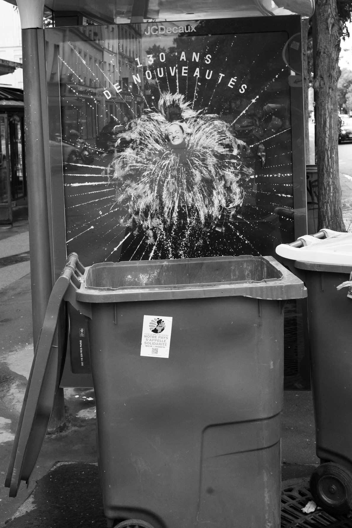 Affiche publicitaire festive célébrant “130 ans de nouveautés” partiellement masquée par une poubelle, créant un contraste entre glamour et ordinaire urbain.