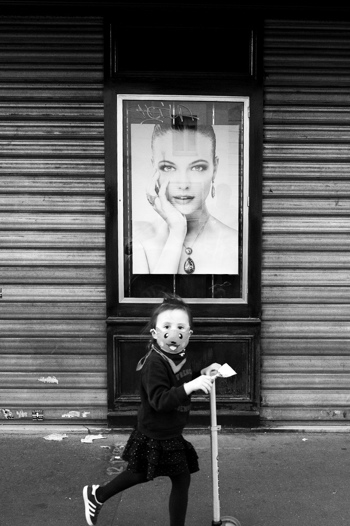 Petite fille en trottinette portant un masque animalier, passant devant une vitrine avec une affiche glamour d'une femme, créant un contraste fort entre les deux figures.