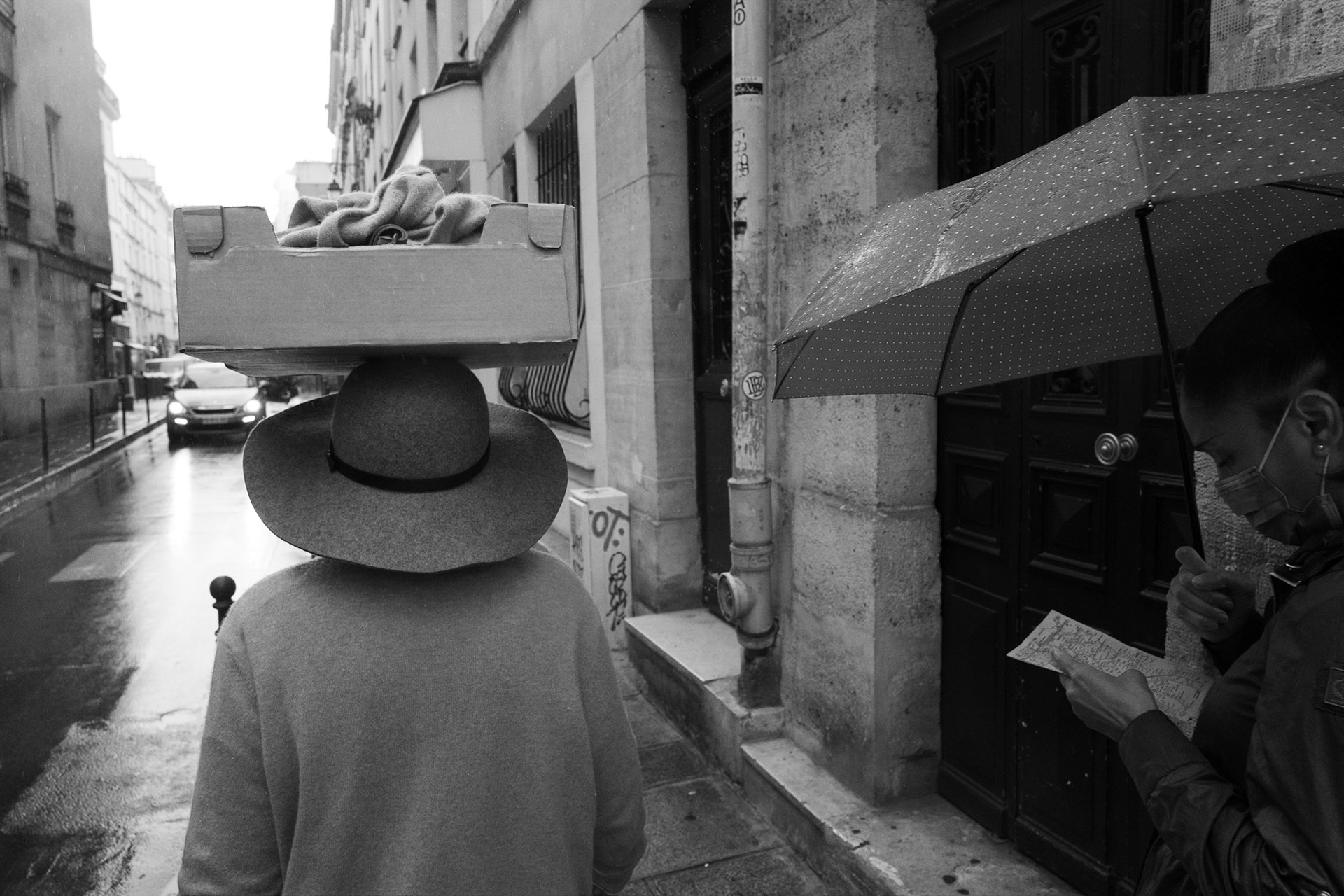 Scène de rue un jour de pluie : une personne vue de dos avec un chapeau large portant une caisse sur la tête, croise une femme sous un parapluie lisant une carte.