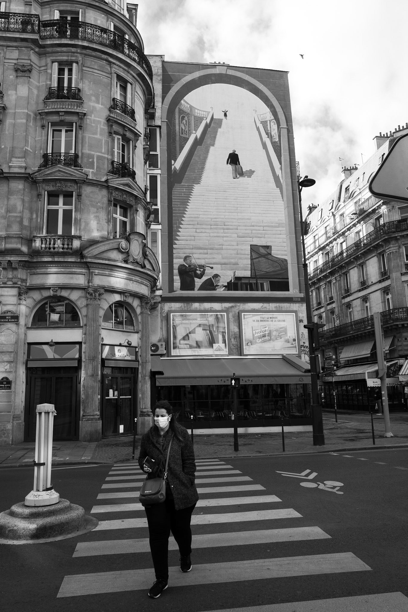 Une femme masquée traverse un passage piéton devant une fresque monumentale représentant un escalier irréel, créant une illusion d’optique entre l'art et l'architecture parisienne.