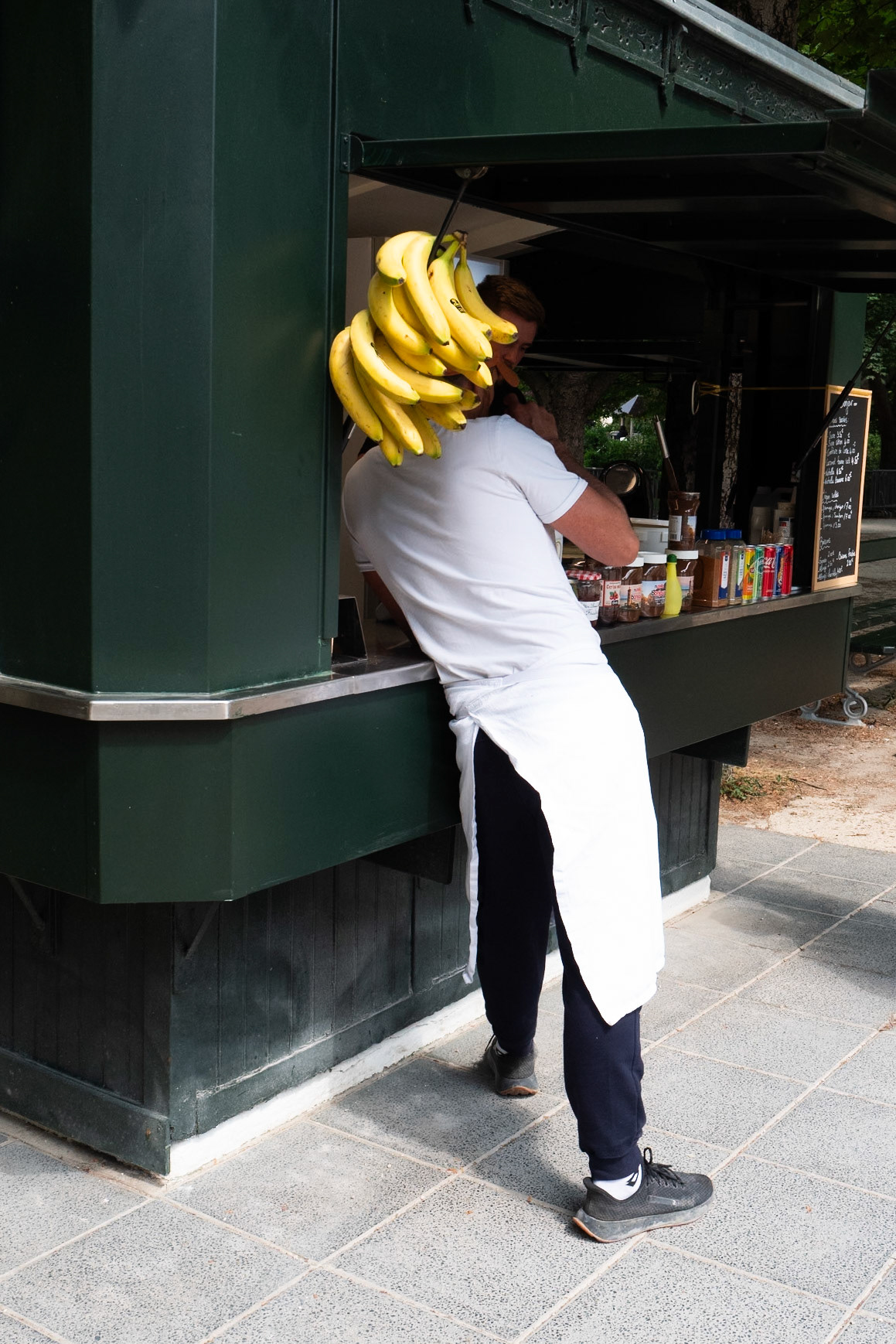 Un vendeur accoudé à un kiosque, le visage en partie caché par une grappe de bananes suspendues, créant un effet visuel décalé comme un chapeau tropical involontaire.