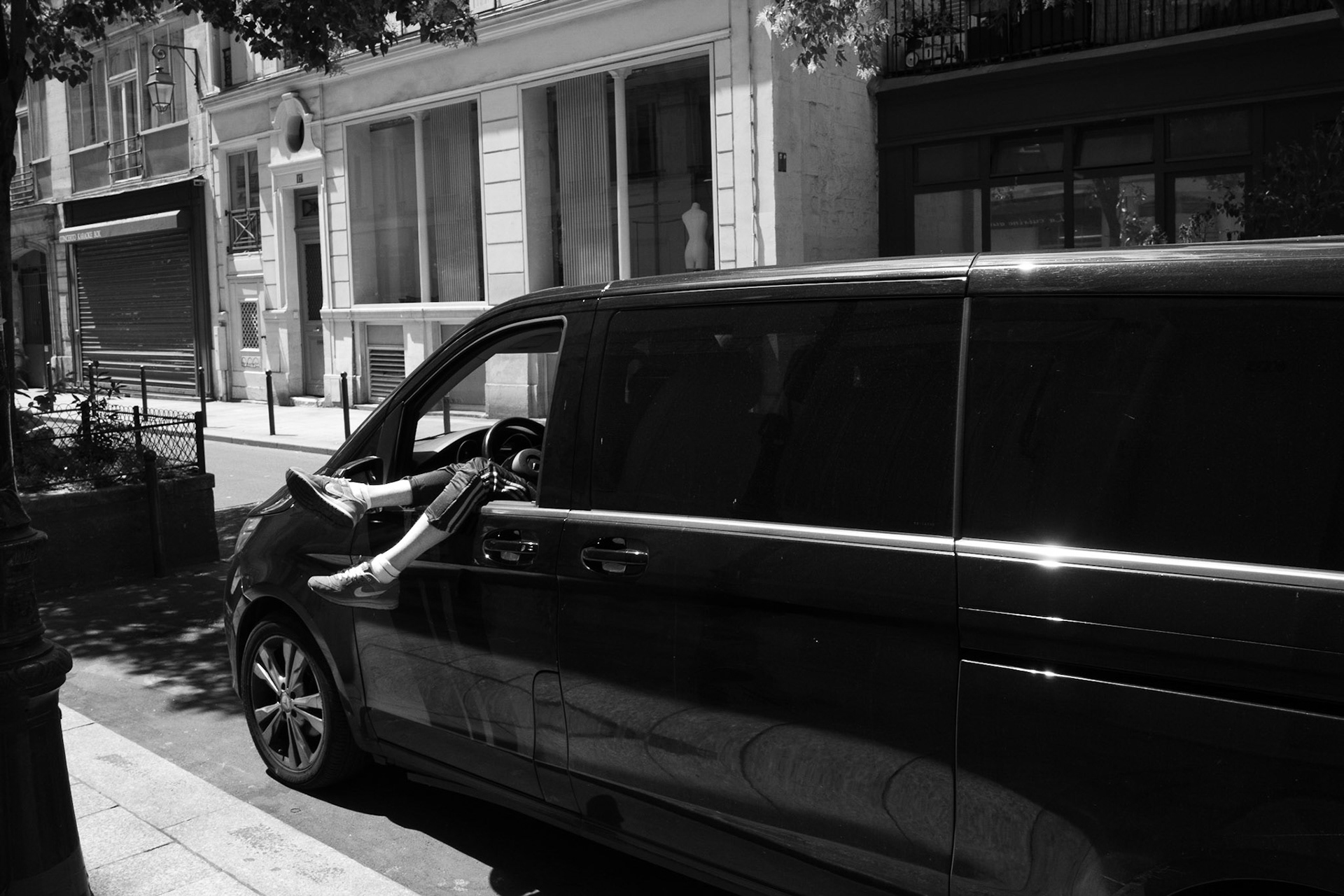 Une paire de jambes sortant de la fenêtre conducteur d’un van noir stationné, les pieds croisés, capturant un instant inattendu et ludique dans la rue.