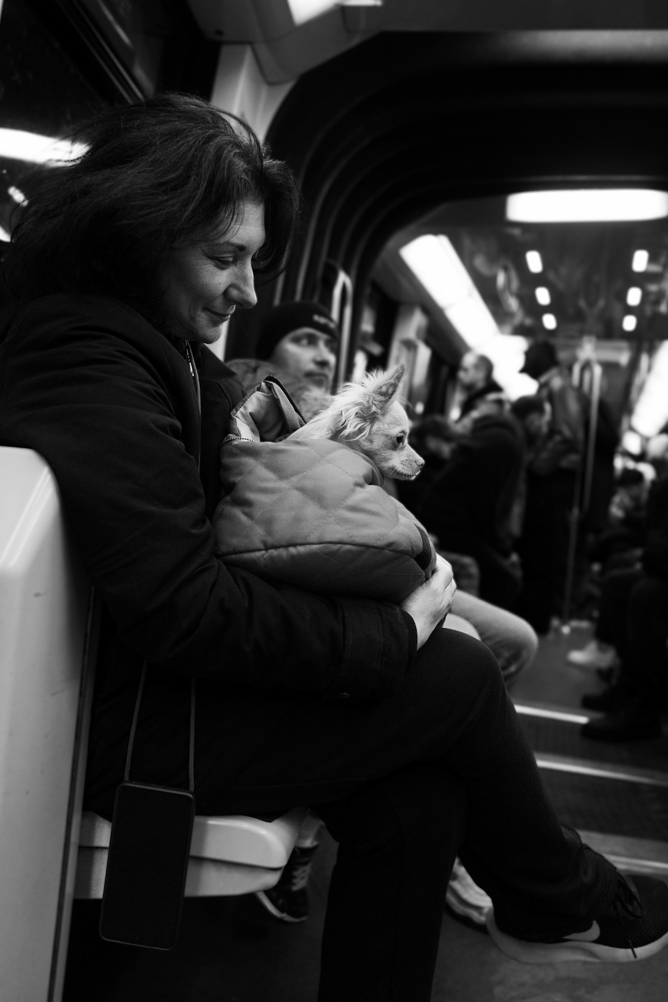 Dans un métro bondé, une femme souriante tient un petit chien enveloppé dans sa veste, le regardant avec tendresse pendant que les passagers derrière restent absorbés dans leurs pensées.