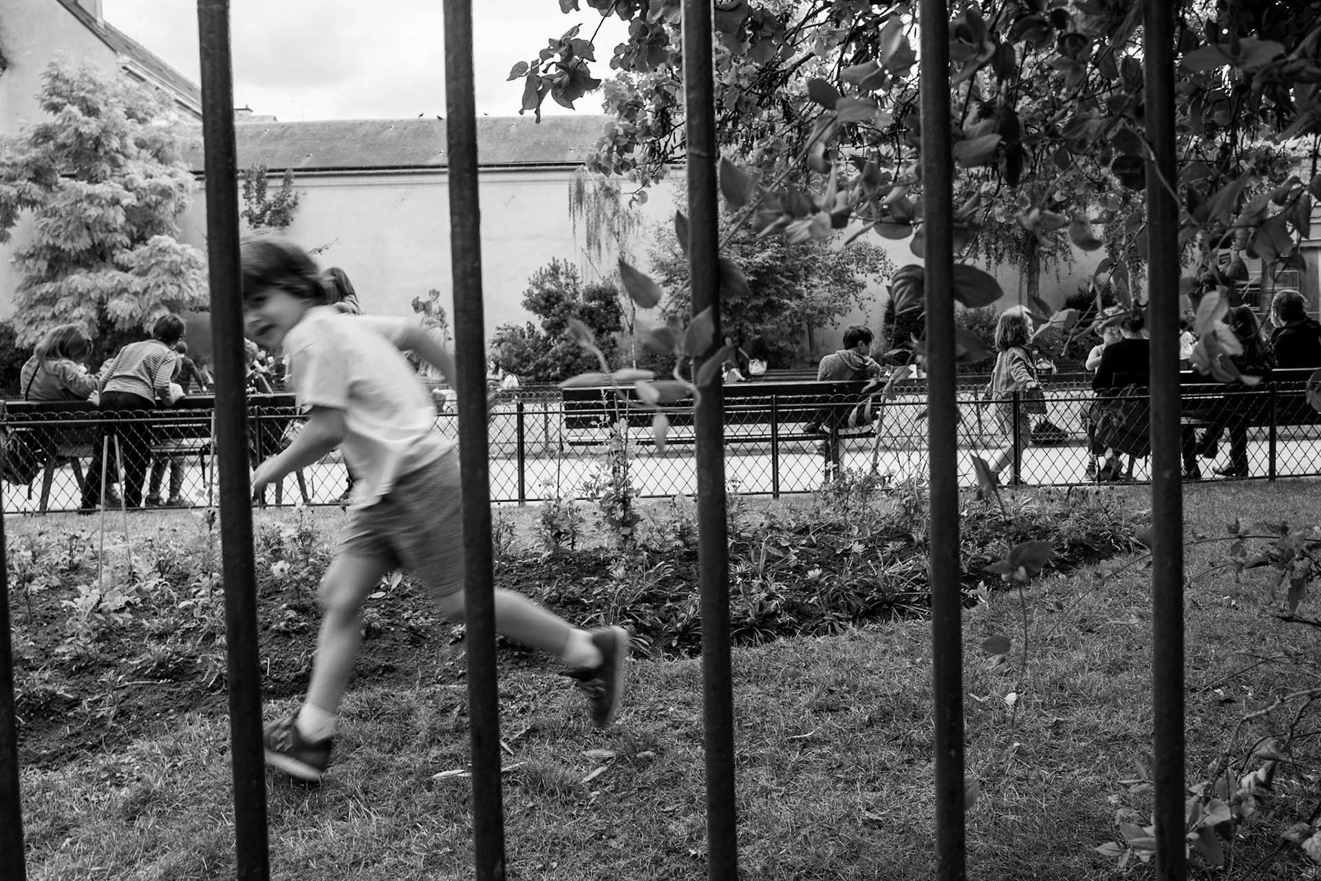 Un enfant court devant des grilles dans un square parisien, flou de mouvement, pendant que d’autres enfants jouent en arrière-plan sur des bancs et autour d’une sculpture.