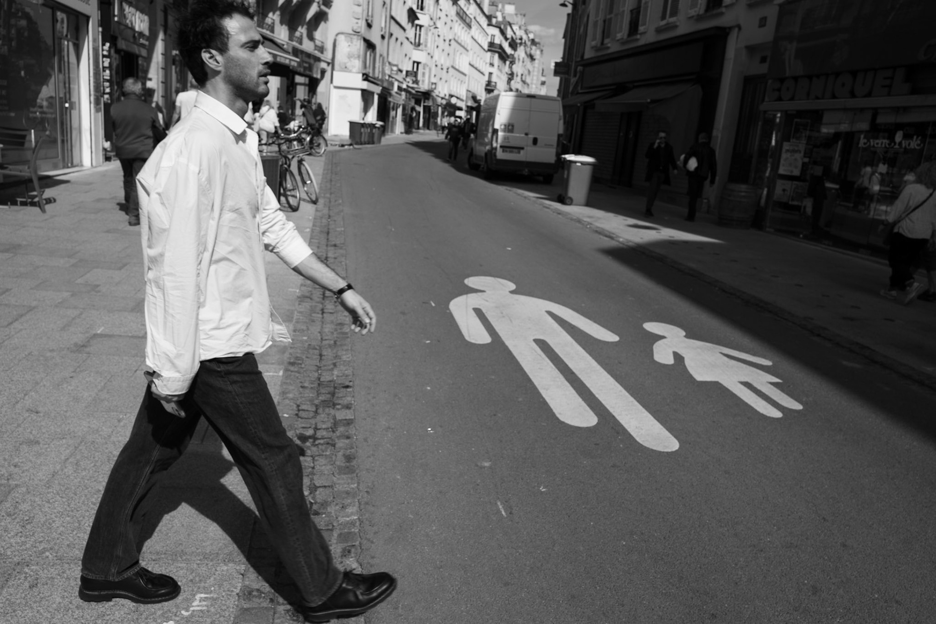 Un homme traverse la rue devant des pictogrammes peints au sol représentant un adulte et un enfant, comme une mise en scène graphique entre le réel et le symbole.