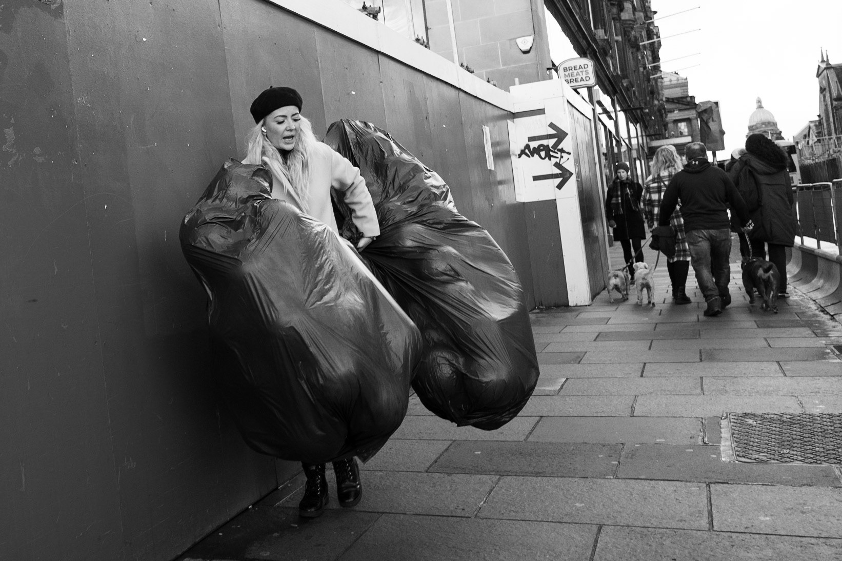 Une femme élégante coiffée d’un béret lutte pour porter deux énormes sacs-poubelles noirs dans une rue commerçante, une scène à la fois absurde et stylée.