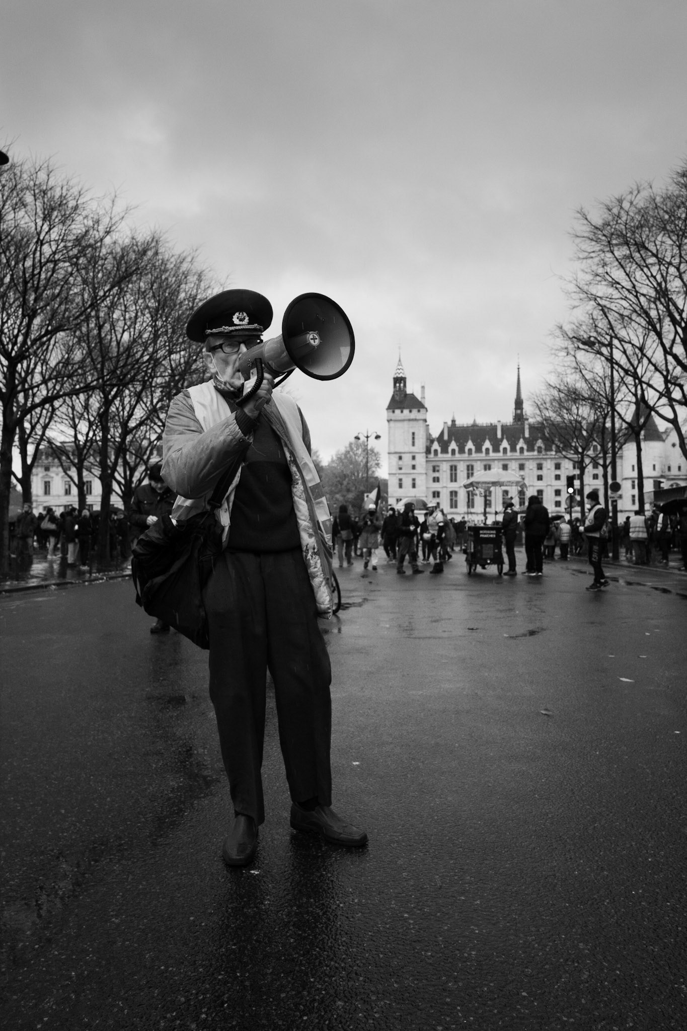 Un homme âgé en uniforme, casquette vissée et mégaphone à la main, harangue une foule lors d’une manifestation, avec la Conciergerie en toile de fond.