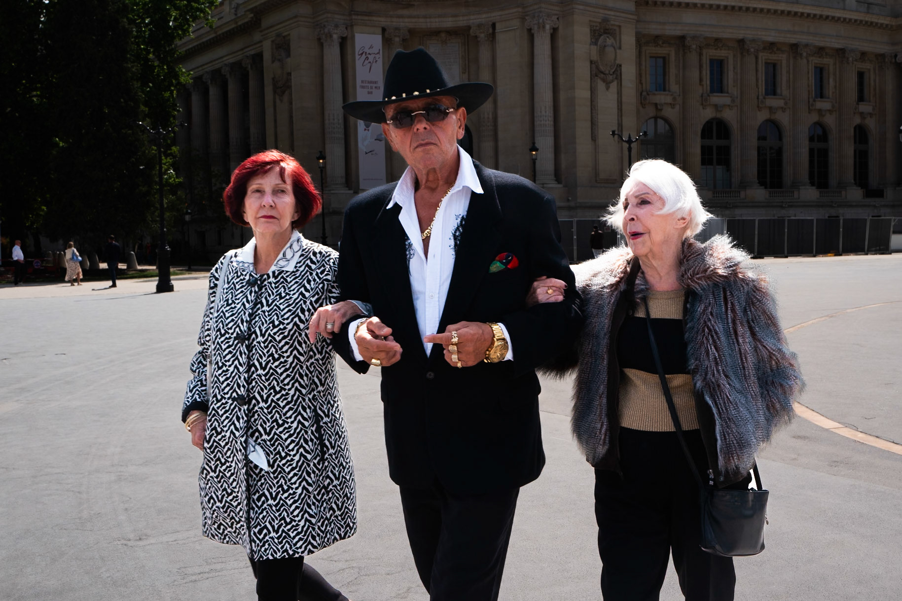 Un homme en costume noir, lunettes de soleil et chapeau, marche bras dessus bras dessous avec deux femmes âgées bien habillées, en pleine lumière devant un bâtiment classique parisien.