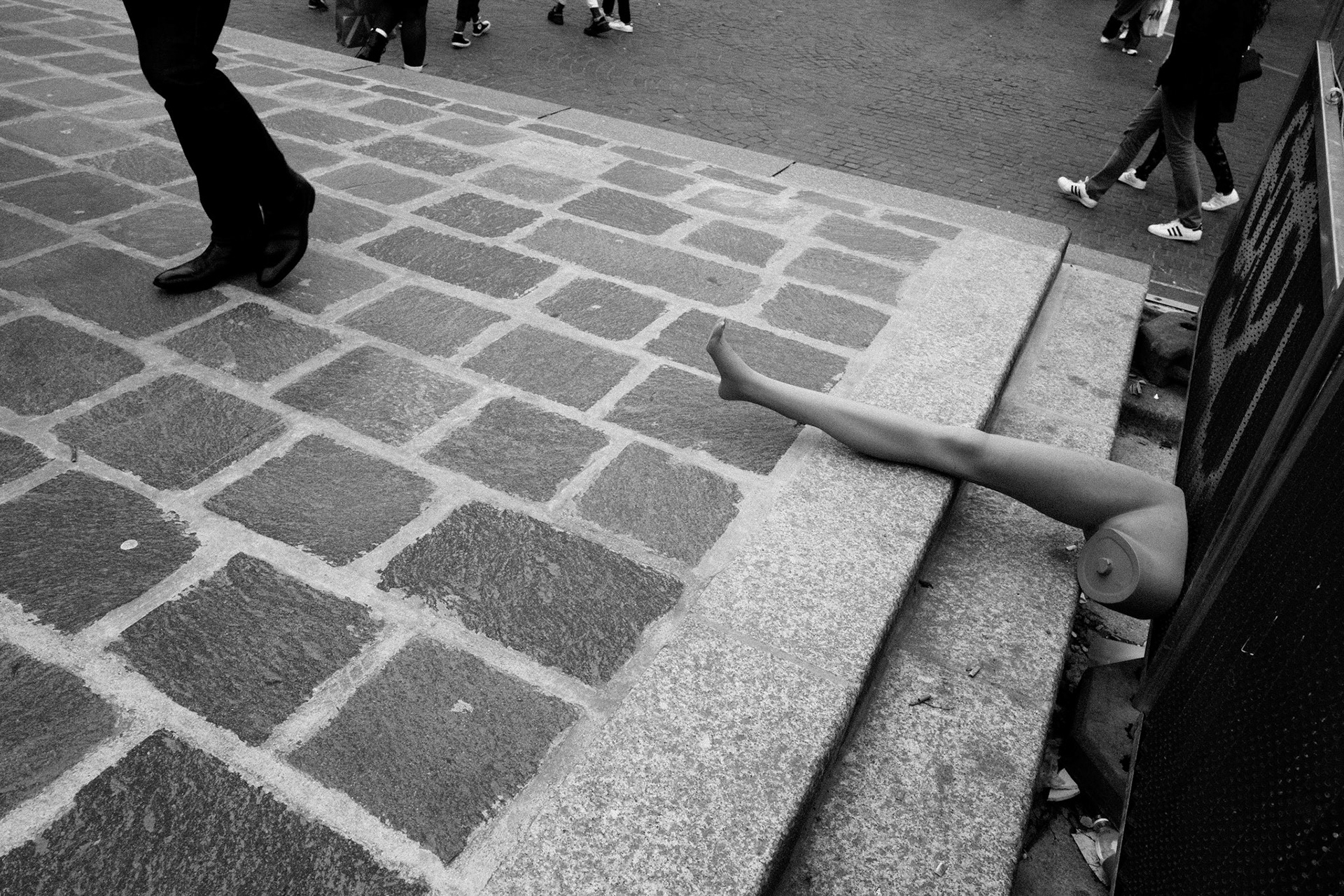 Jambe de mannequin abandonnée sortant d’une bouche d’égout sur un trottoir pavé, avec des passants au second plan. Une scène à la fois absurde et graphique.