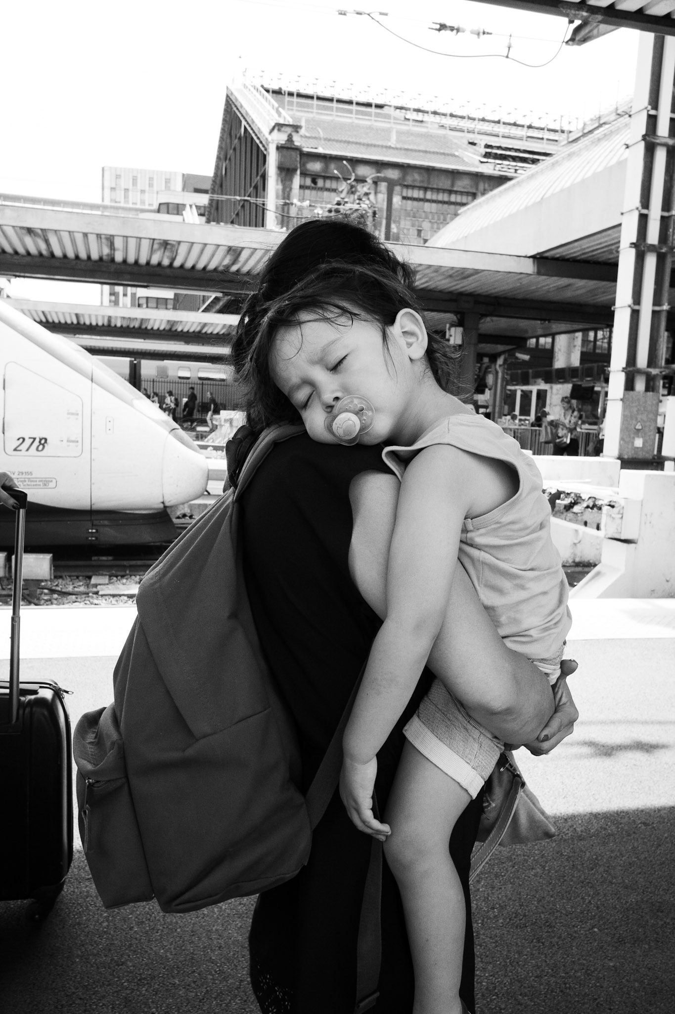 Une petite fille endormie avec une tétine est portée dans les bras sur un quai de gare, blottie contre l’épaule d’un adulte, un TGV en arrière-plan.