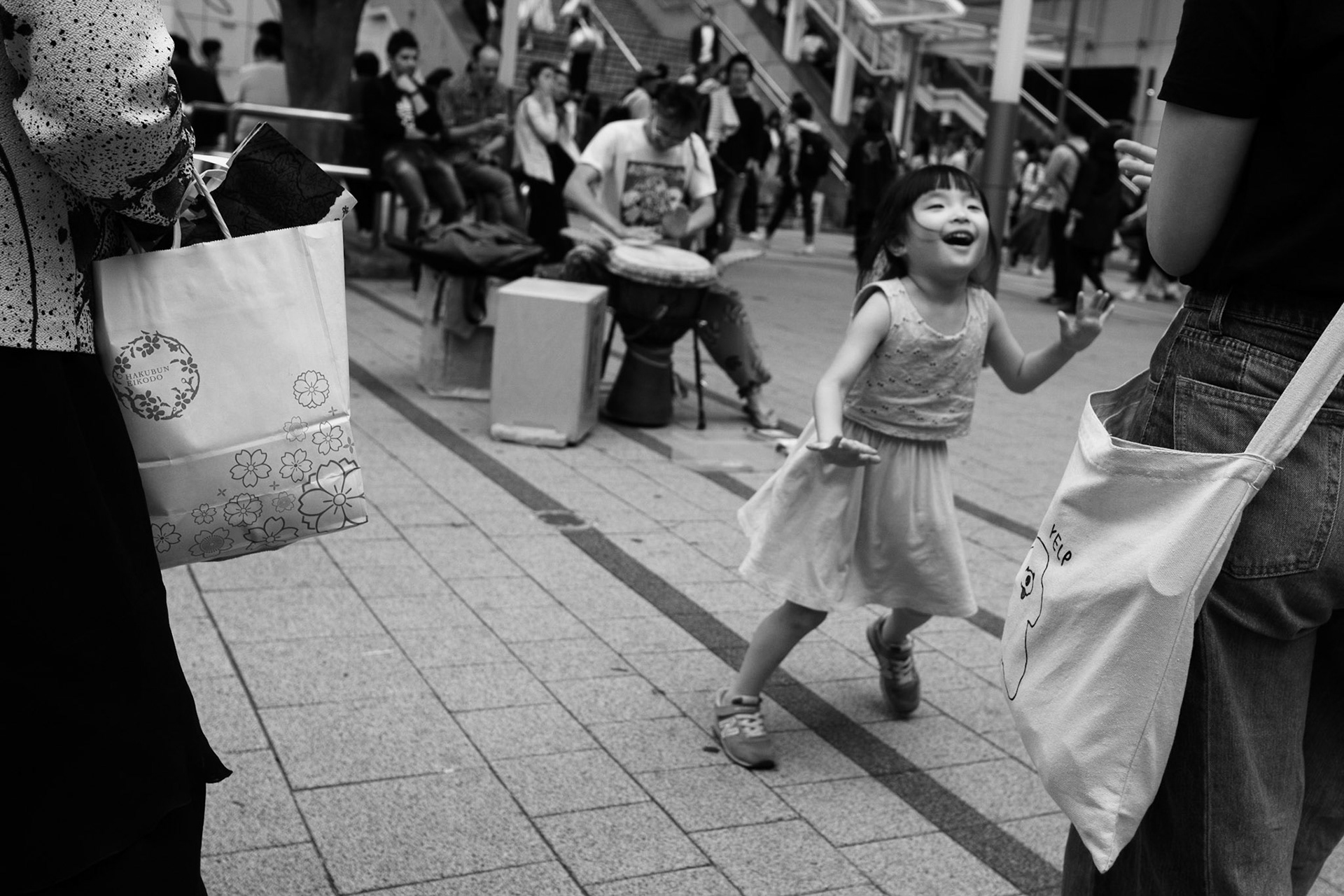 Petite fille en robe légère courant joyeusement entre deux adultes dans une rue piétonne animée, alors qu’un musicien joue du djembé au second plan, créant une scène vivante et spontanée.
