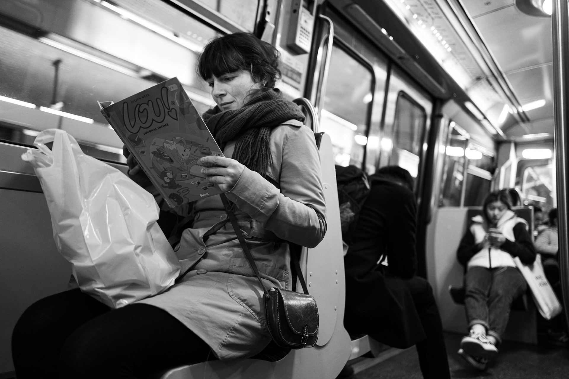 Une femme lit une bande dessinée intitulée « Lou ! » dans une rame de métro parisien. Assise seule sur son siège, elle semble absorbée, tandis que les autres passagers regardent leur téléphone ou fixent le vide.
