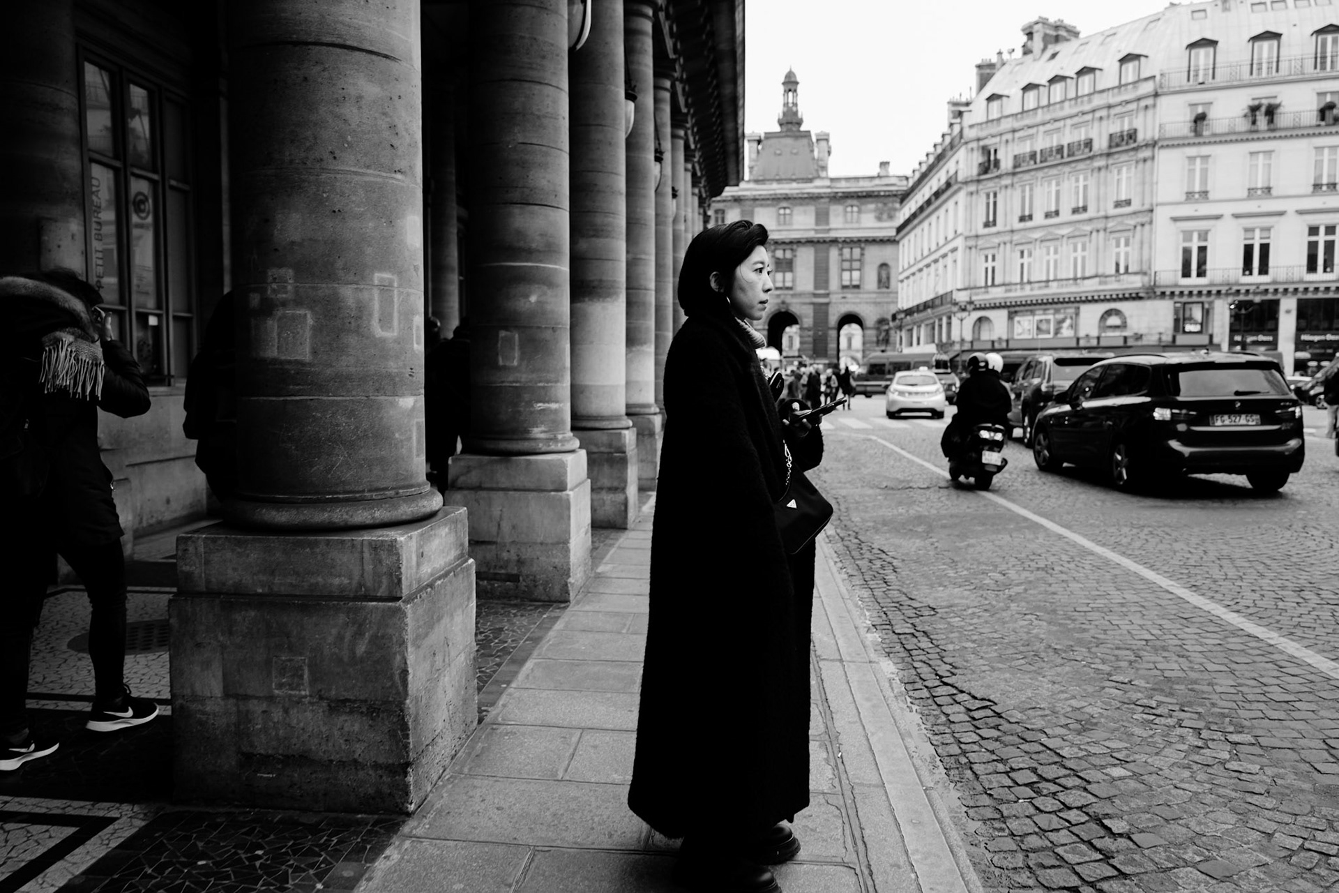 Une femme vêtue de noir attend sur un trottoir sous les arcades du Palais Royal, le regard tourné vers la circulation. À l’arrière-plan, l’architecture haussmannienne encadre la scène avec sobriété.