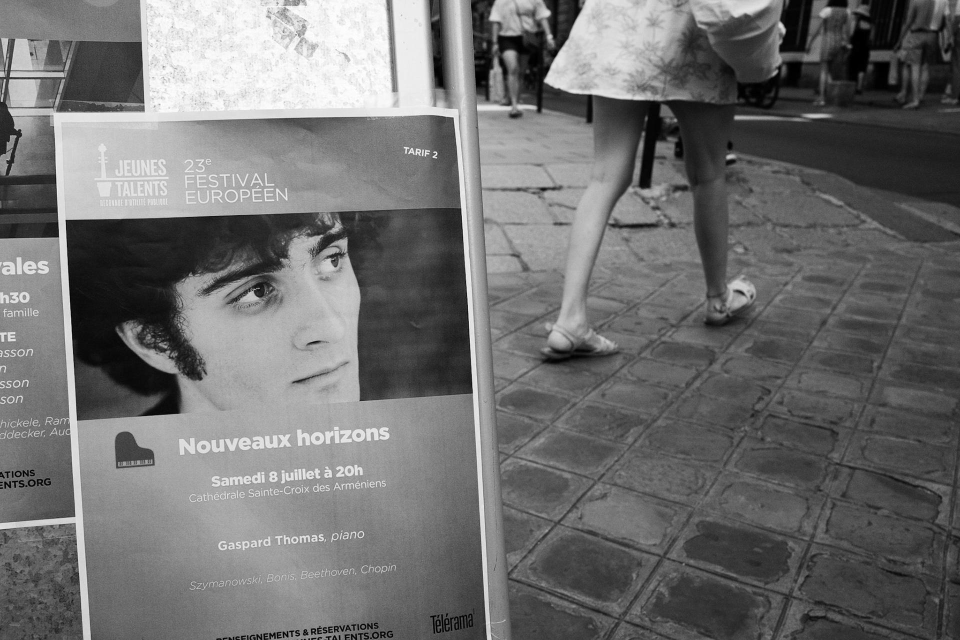 Affiche d’un festival montrant un homme qui regarde à côté du passage vif d’une jeune femme en robe, jambes en mouvement sur le pavé.