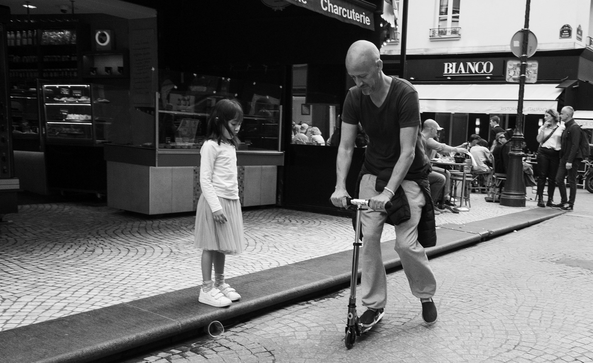 Un homme glisse sur une trottinette sous le regard d’une fillette debout sur une bordure, captés dans une rue pavée animée d’un quartier commerçant.