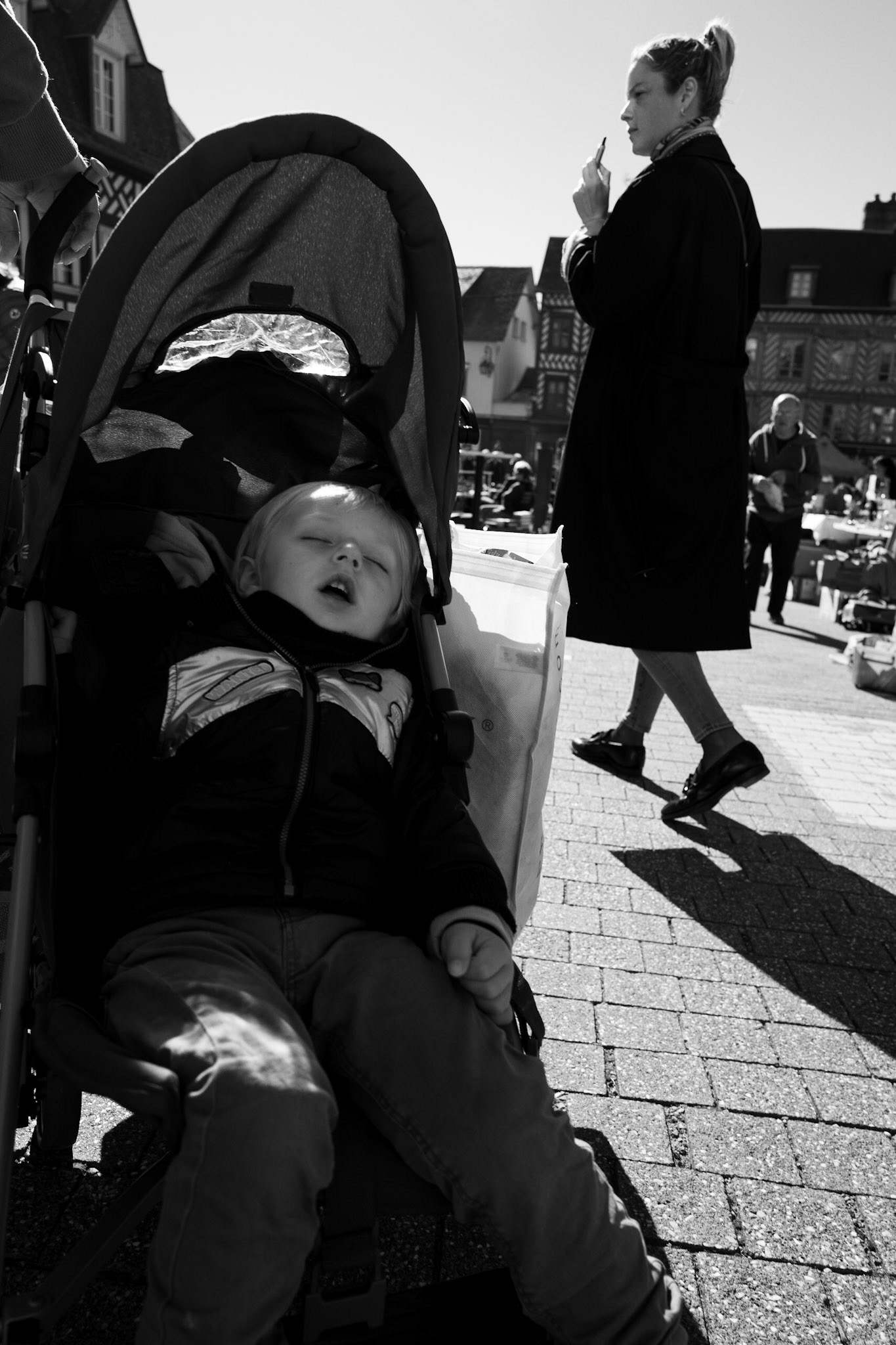 Un jeune enfant endormi dans une poussette au premier plan, pendant qu’une femme passe derrière, absorbée par son téléphone, dans une scène de marché baignée de lumière.