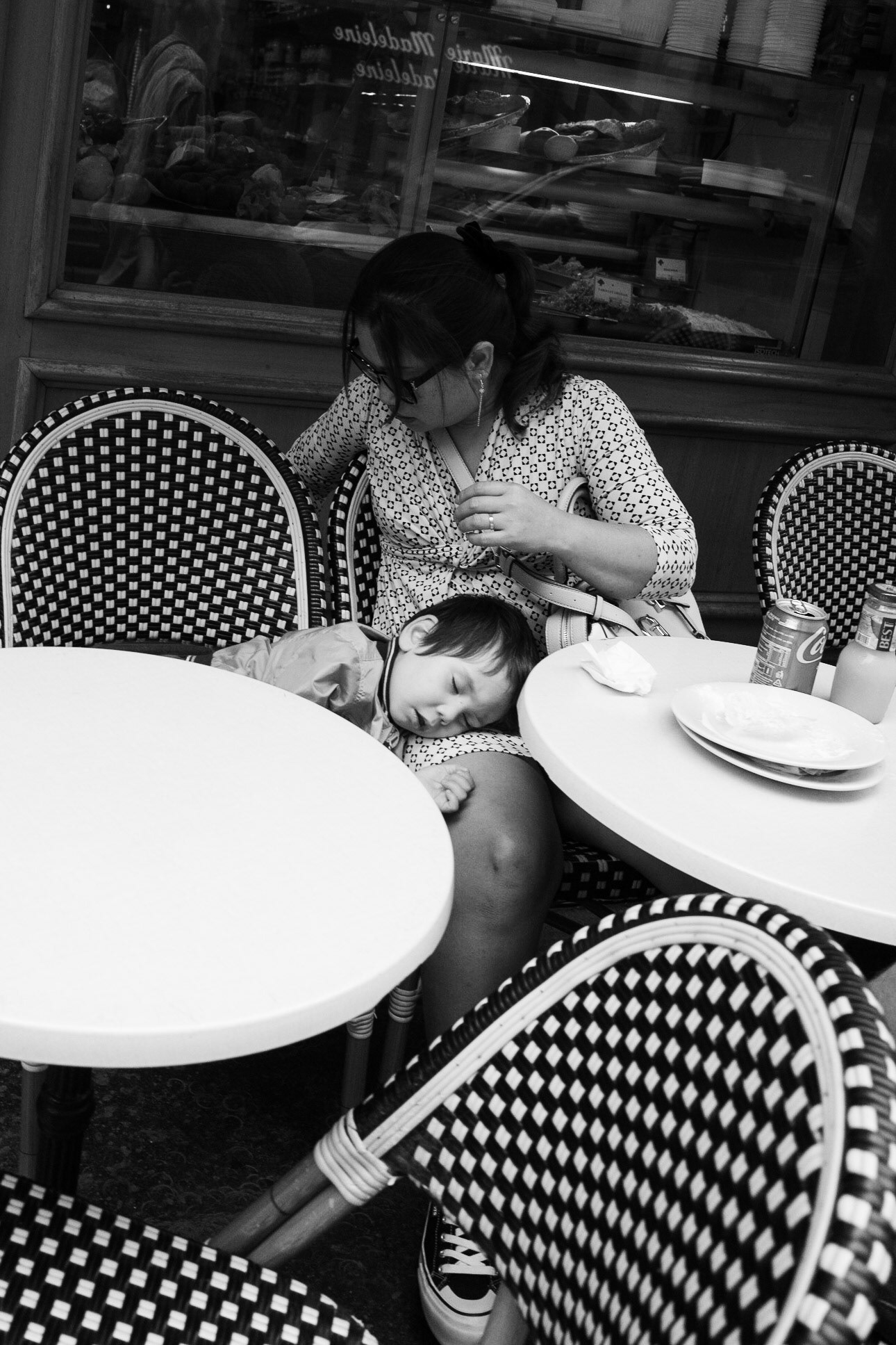 Scène de terrasse parisienne : un enfant dort allongé sur les genoux de sa mère, entre des chaises bistro et des tables vides, après un repas.