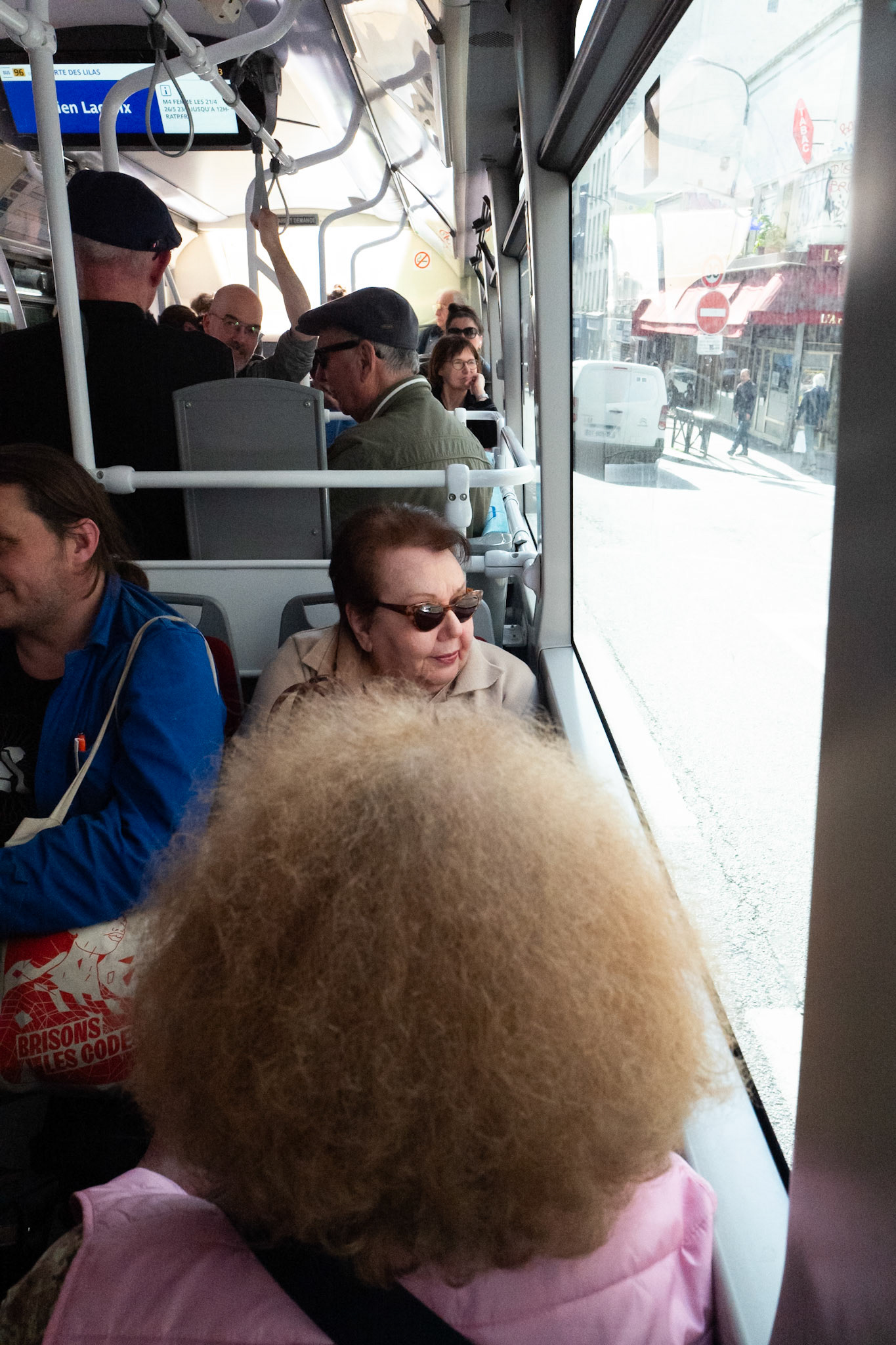 Scène de bus parisien en journée : les passagers assis et debout se croisent du regard ou contemplent la rue, tandis qu’une femme au premier plan domine la composition avec sa chevelure volumineuse.