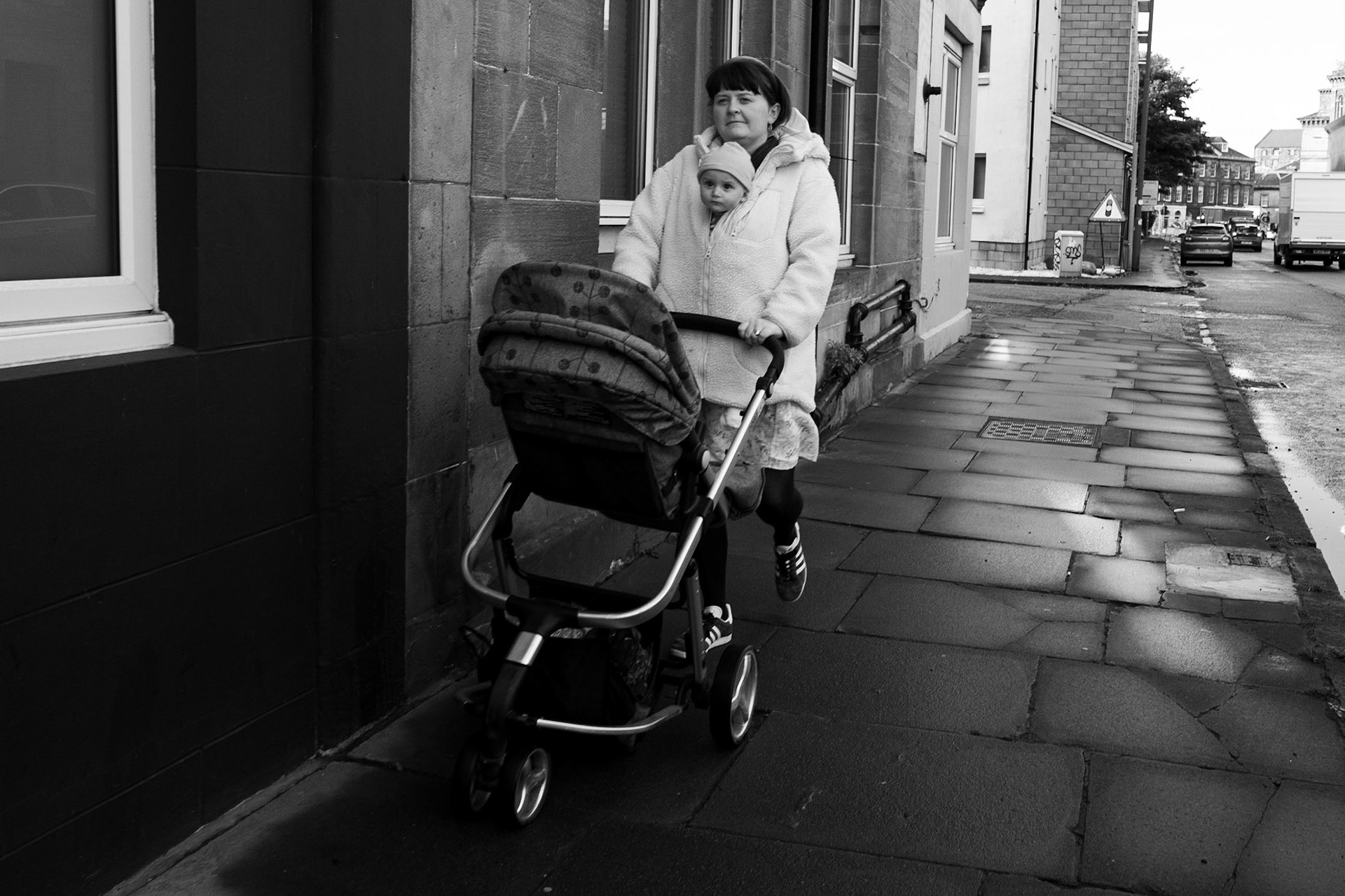 Une femme pousse une poussette sur un trottoir humide, l’enfant bien emmitouflé dans une couverture. Les pavés brillent sous la lumière diffuse.