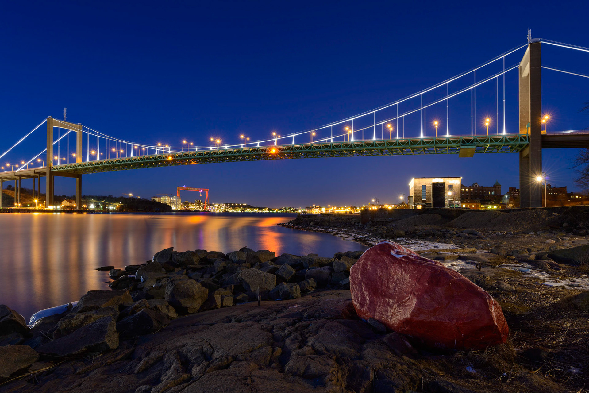 Illuminated bridge over a river with a red boulder in the foreground