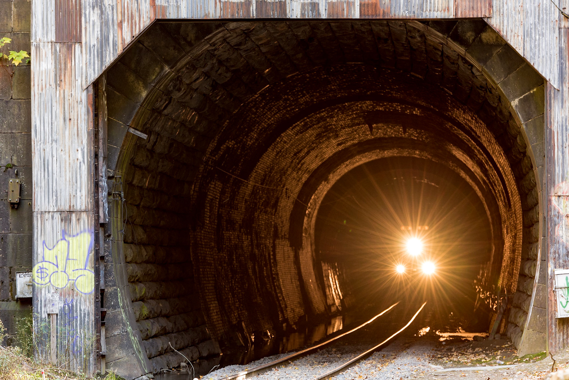 We were planning to photograph the east portal of the historic and infamous Hoosac Tunnel (known as the Bloody Pit after its construction) when a native of North Adams told us how to get to the west portal. The west portal has a rather ugly winter weather door framing it now, but we decided to take the side trip As we approached the portal, several people were wandering around the tunnel entrance and going a few feet inside. Looking down the tunnel, however, I saw a dim light well down the tunnel. At first, I thought it was someone crazy enough to walk back into the tunnel. I soon realized, however, that the light was slowly resolving into the signature three-light triangle of a locomotive, and I could hear the echo of air brakes. I quickly set up my tripod and started to focus on the light. Because of the lack of contrast in the tunnel, I had to focus manually, and then pull focus as the train slowly made its way toward me. This shot was the best of the group, in my opinion, because the illuminated interior of the tunnel shows a richness of detail.Date: 10 October 2016Location: North Adams, Massachusetts, United StatesOriginal resolution: 36 MPProcessing: Processed from RAW using Adobe Photoshop Lightroom CC 2015