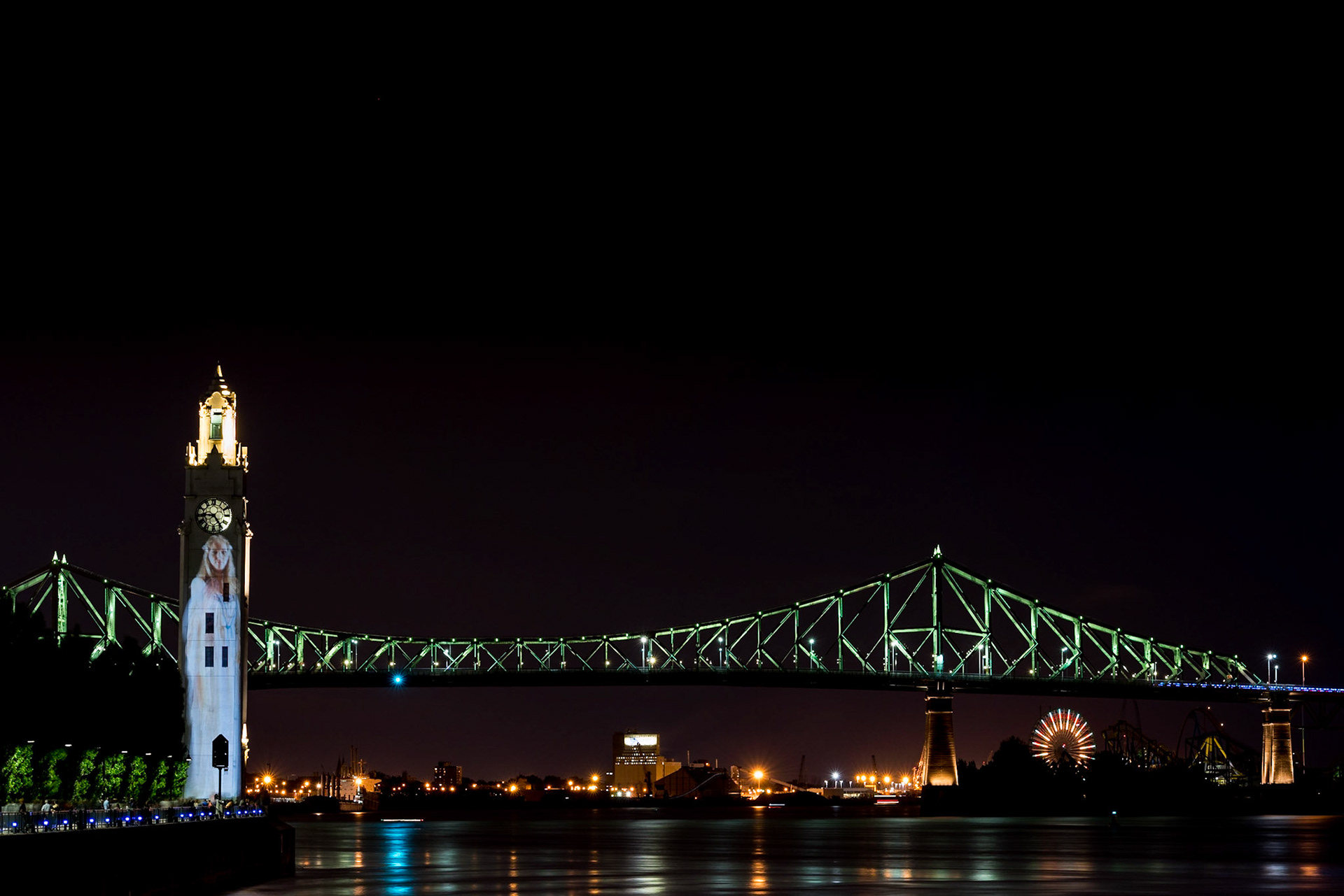 I shot this image of the Montréal waterfront while waiting for the fireworks to begin. The Saint Lawrence River passes under the Jacques Cartier bridge. The ferris wheel of the La Ronde amusement park, site of the fireworks display, is visible under the right side of the bridge.On the left, an image of a woman is clearly visible on the Clock Tower. The projection is part of a citywide art installation known as Cité Mémoire. The woman represents Suzanne, of the 1966 Leonard Cohen song, which is perhaps his most famous.Date: 26 July 2017Location: Montréal, Québec, CanadaOriginal resolution: 36 MPProcessing: Processed from RAW using Adobe Photoshop Lightroom 6