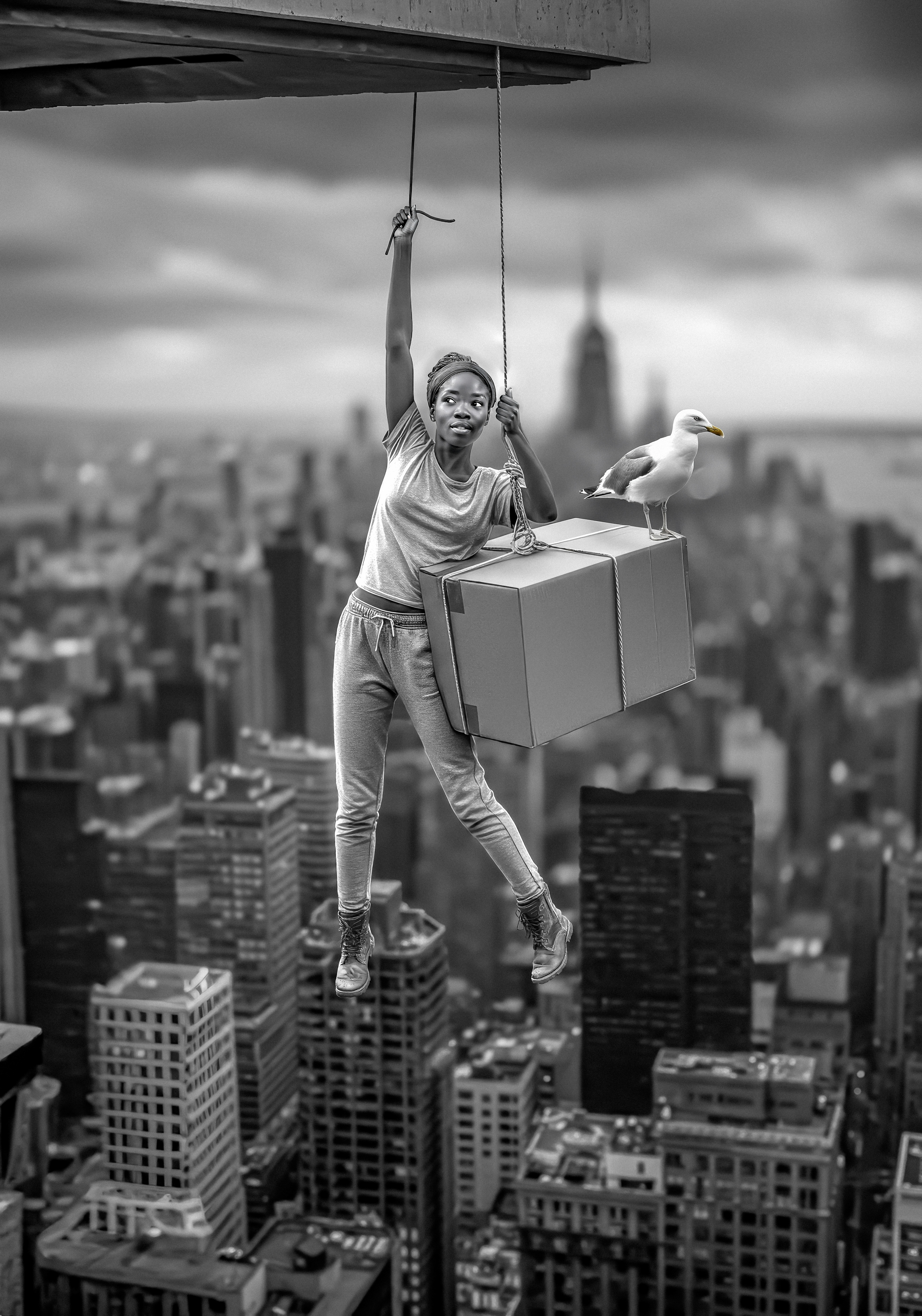 "In a striking black-and-white composition, Bob Orsillo explores the metaphor of life's choices through surreal imagery. A lone figure dangles precariously from a rope high above an expansive cityscape, holding a box that symbolizes the weight and complexity of decisions. A seagull perched atop the box adds a whimsical touch, reflecting the spontaneity and unpredictability of choices. This thought-provoking photograph merges artistic brilliance with profound symbolic depth."