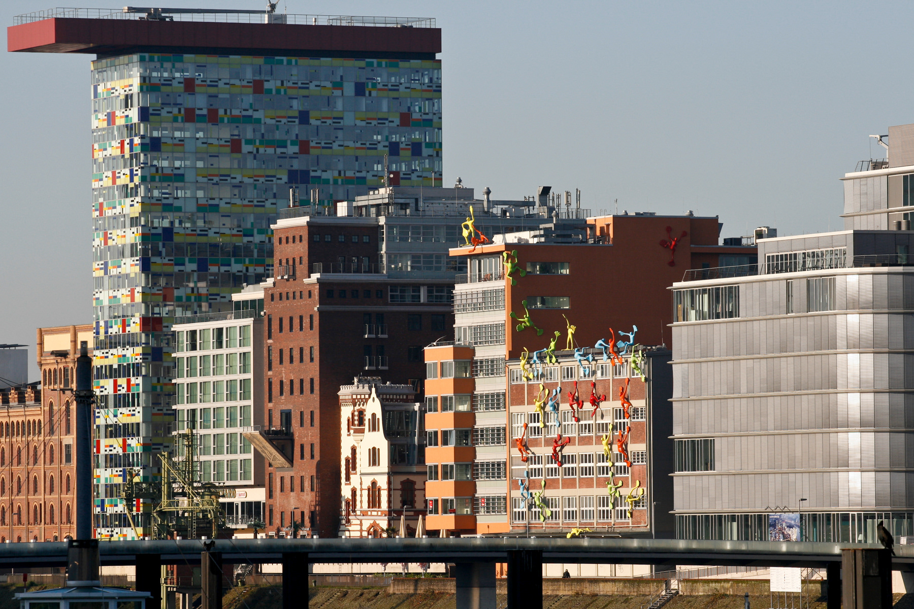 Medienhafen urban development area, November 2009. Or, how an old Rhine harbour has been transformed into a cotemporary place, part by part. As seen from left to right:  Alte Mältzerei (former malthouse of Dortmunder Union Kronen-Braurerei and monument since 1897, redesigned by Bob Gansfort); Colorium (tall building designed by William Alsop); Courtyard Marriot Hotel Düsseldorf Hafen; former silo of the Lamers company renovated into an office building; Kleine Villa (now office building); Neuwerk office building; Roggendorf-Haus (former Roggendorf storage building, renovated by design of Norbert Winkels and featuring the Flossis men from artist Rosalie; Speditionsstrasse 17 by Ingenhoven Overdiek Architects. The Living Bridge provides a 150 m pedestrian and bicycle water crossing.