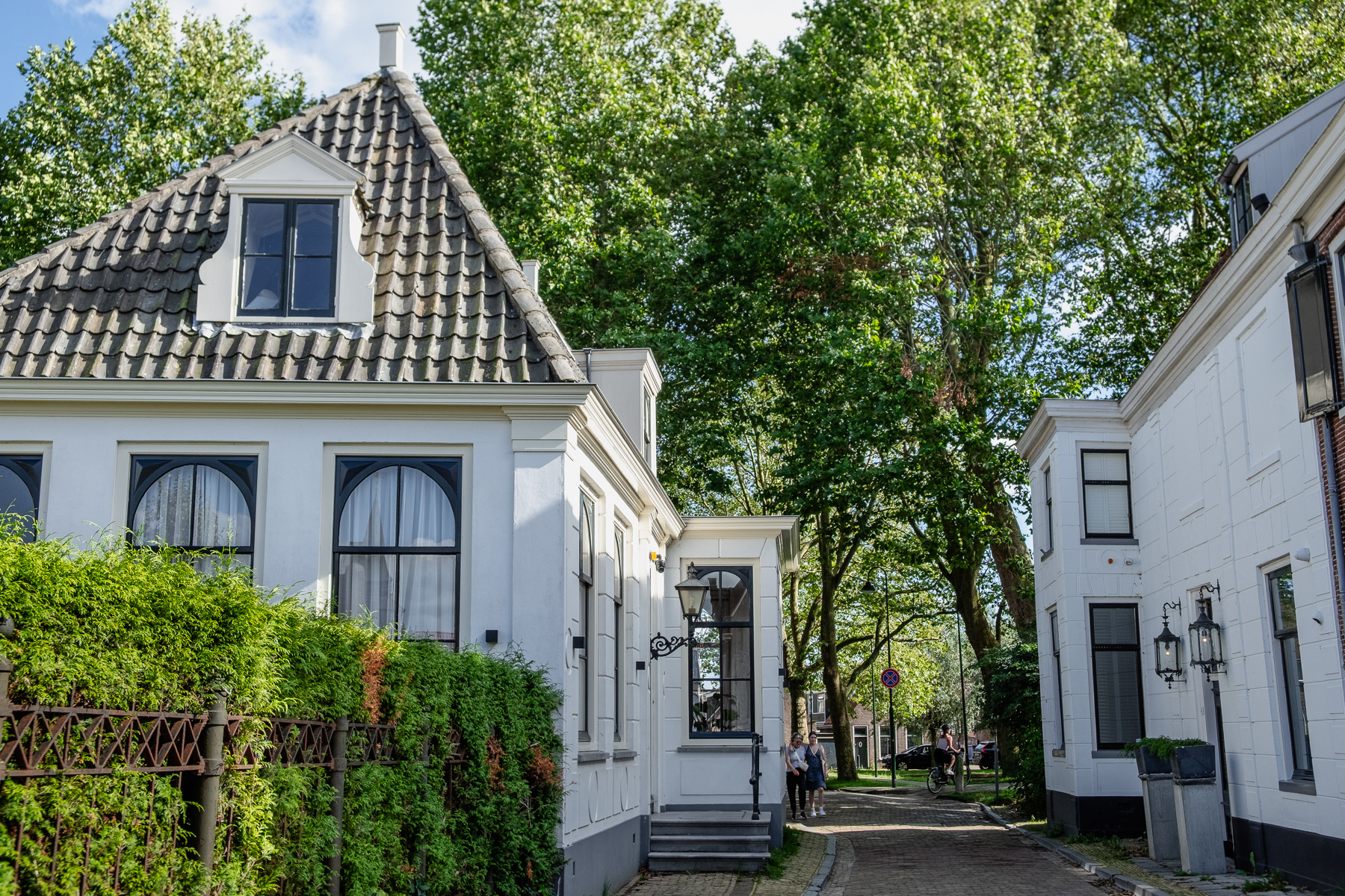 Inside Edam, August 2023. Two hosues and a narrow streat with pedestrians and a cyclist. Trees are towering over all. August 2023.
