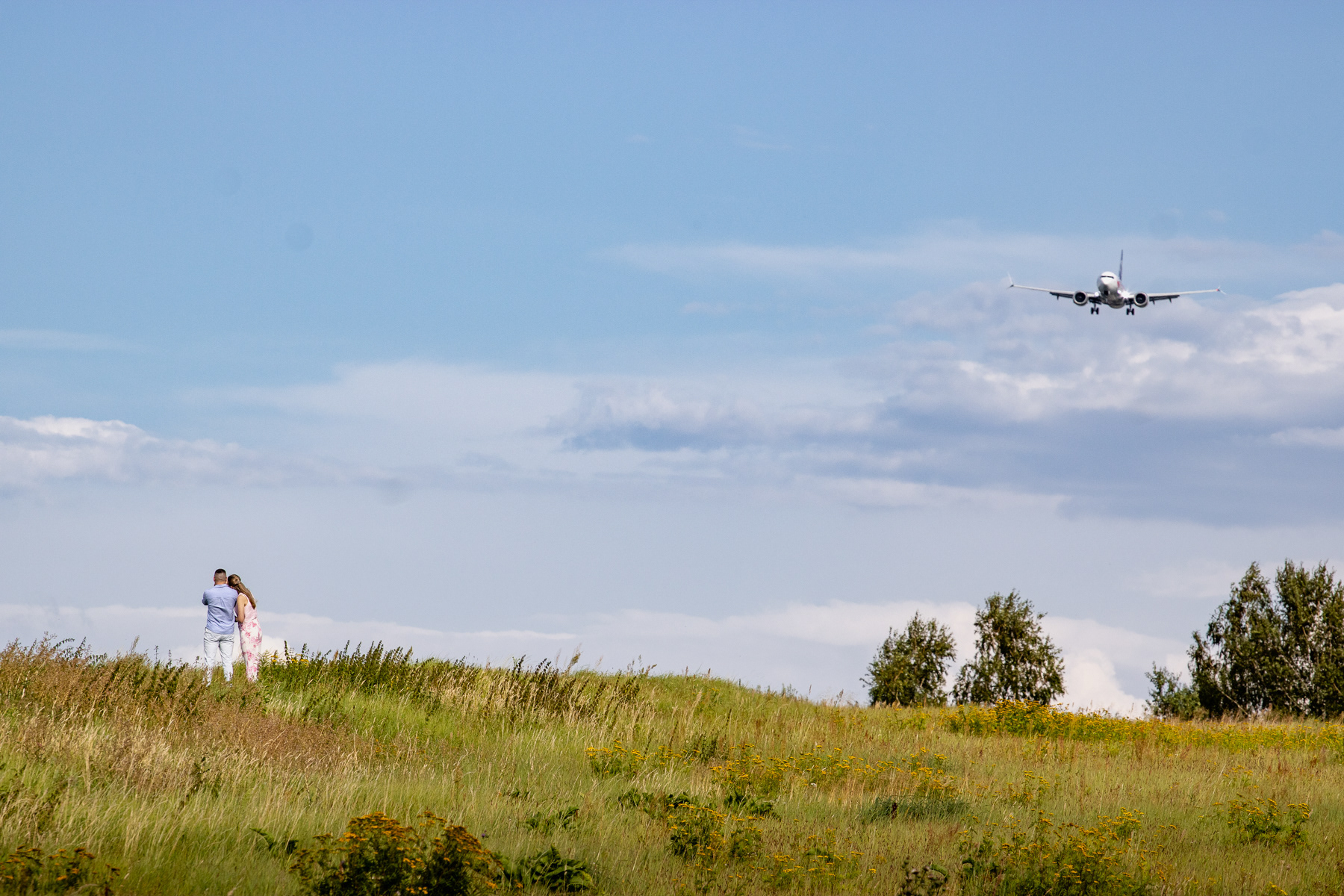 A couple watches a LOT Polish Airlines Boeing 737 Max 8 on final approach to Kraków-Balice Airport, August 2025.
