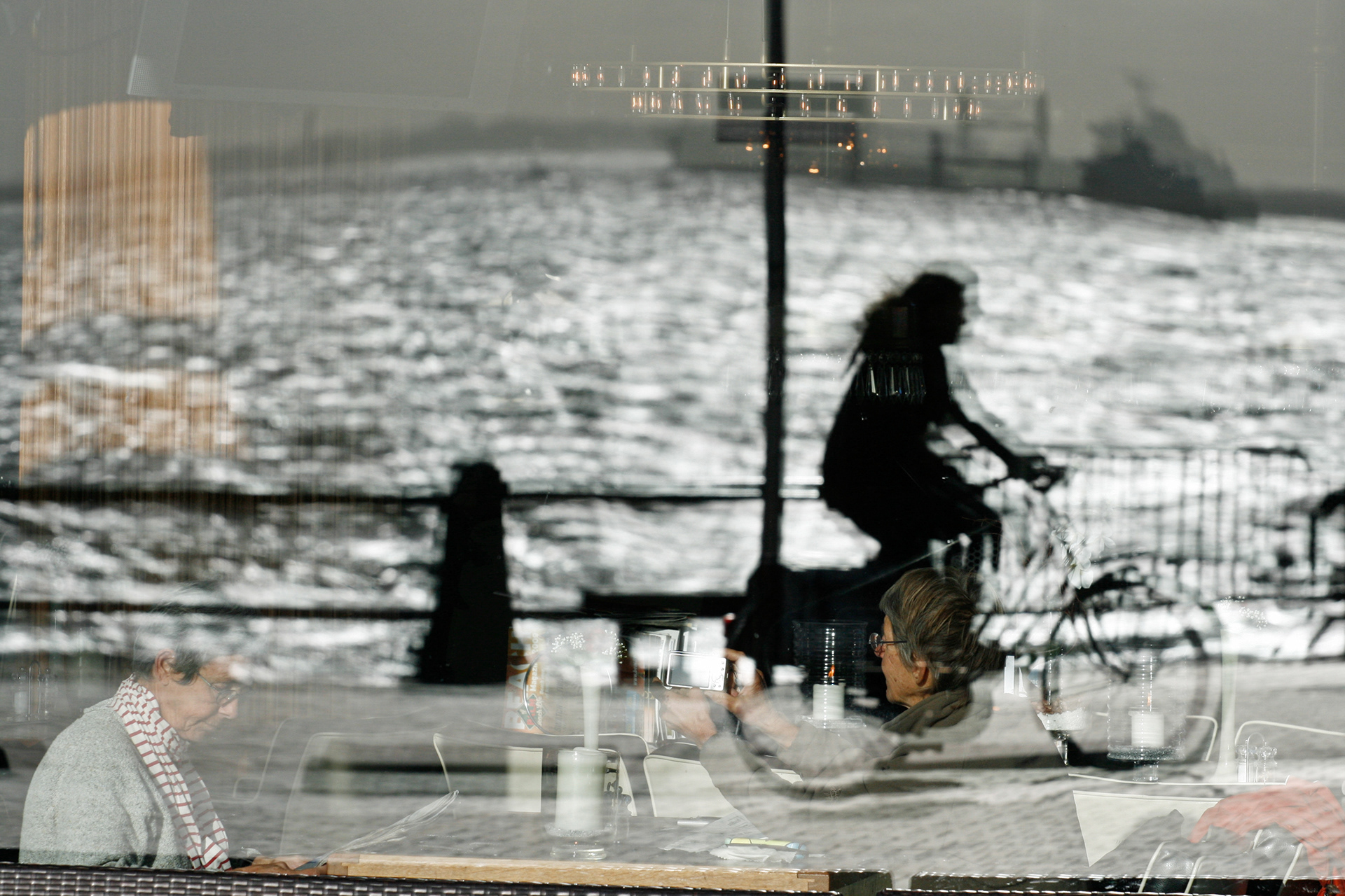 The reflection of a female cyclist and the Westerschelde in a café window. Behind the window two elderly ladies are visible.