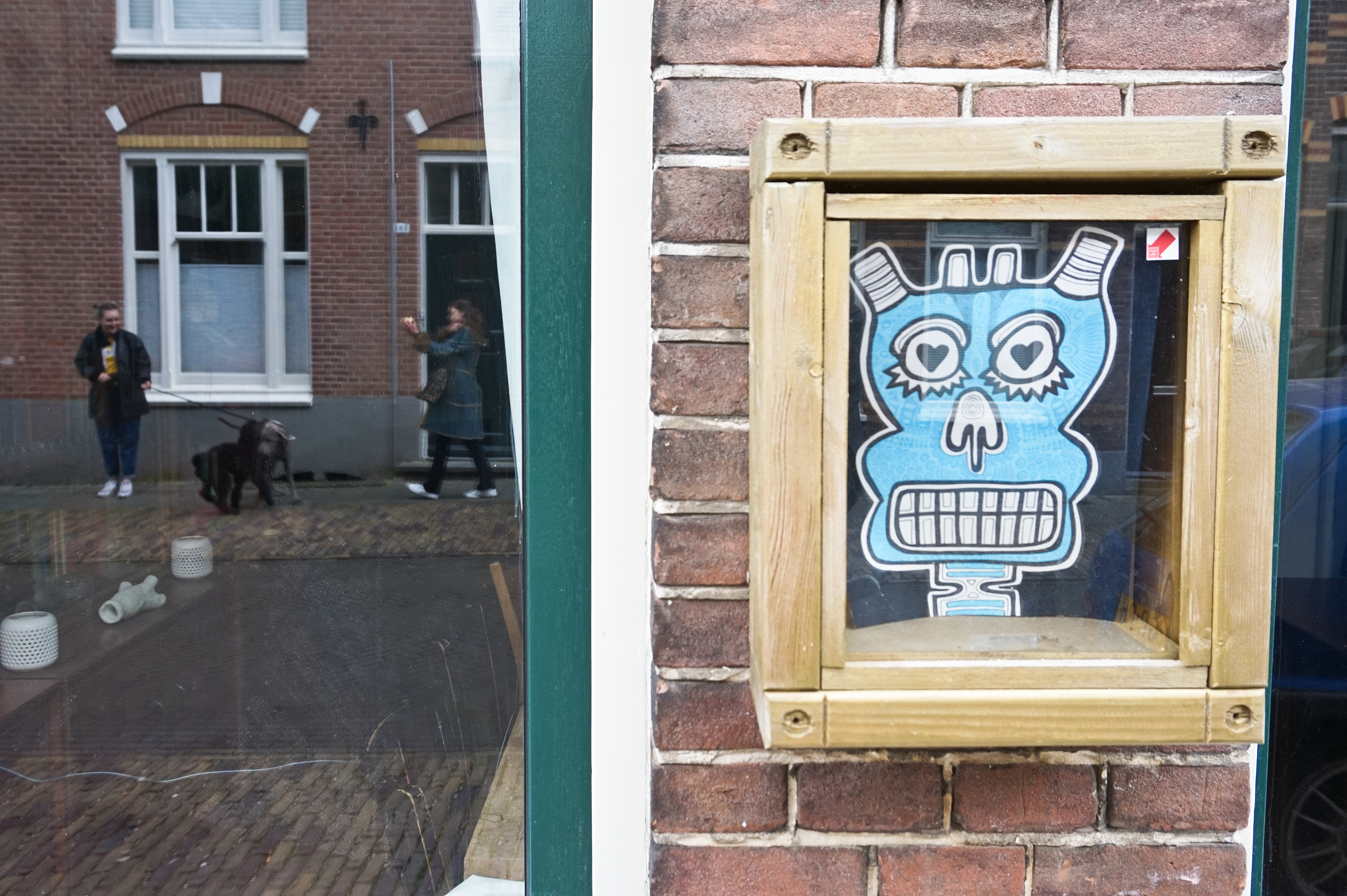An art box on the outer wall. In the reflections of the house window two ladies and a dog.