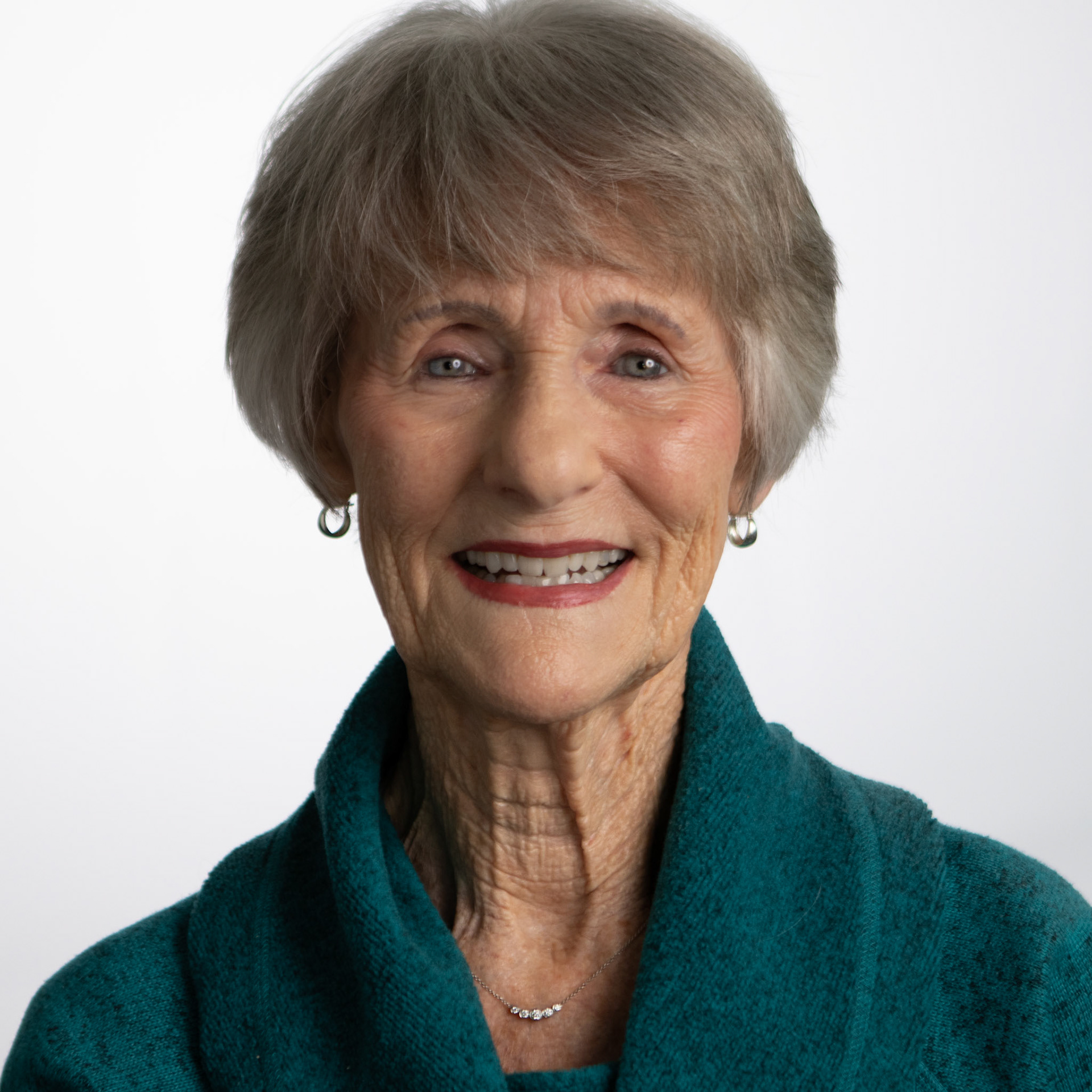 A headshot of a senior woman with short grey hair, smiling with her teeth. She is wearing a teal-colored sweater and a silver necklace with a small pendant. The background is a plain white studio wall.