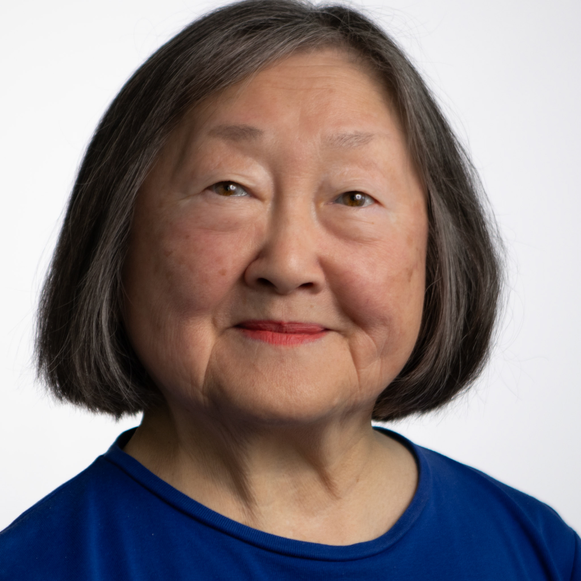 A headshot of an elderly Asian woman with short grey hair and a gentle smile. She is wearing a bright blue shirt and looking directly at the camera.